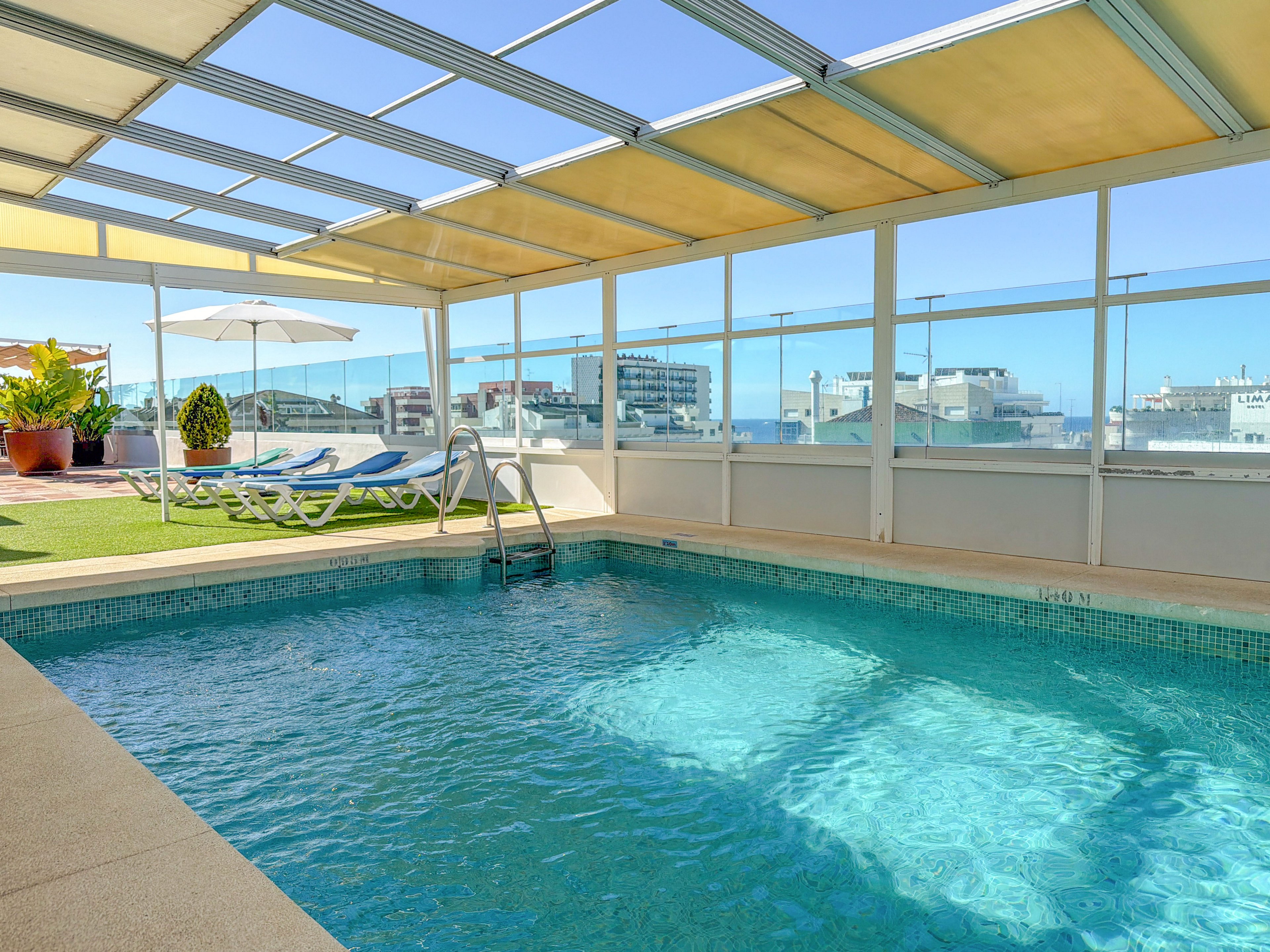 A clear blue rooftop swimming pool is shown under a partial canopy structure. Metal handrails lead into the water, surrounded by a tiled deck. In the background, sun loungers, potted plants, and a large white umbrella sit on a strip of artificial grass. Glass panels frame views of nearby city buildings under a bright, sunny sky.