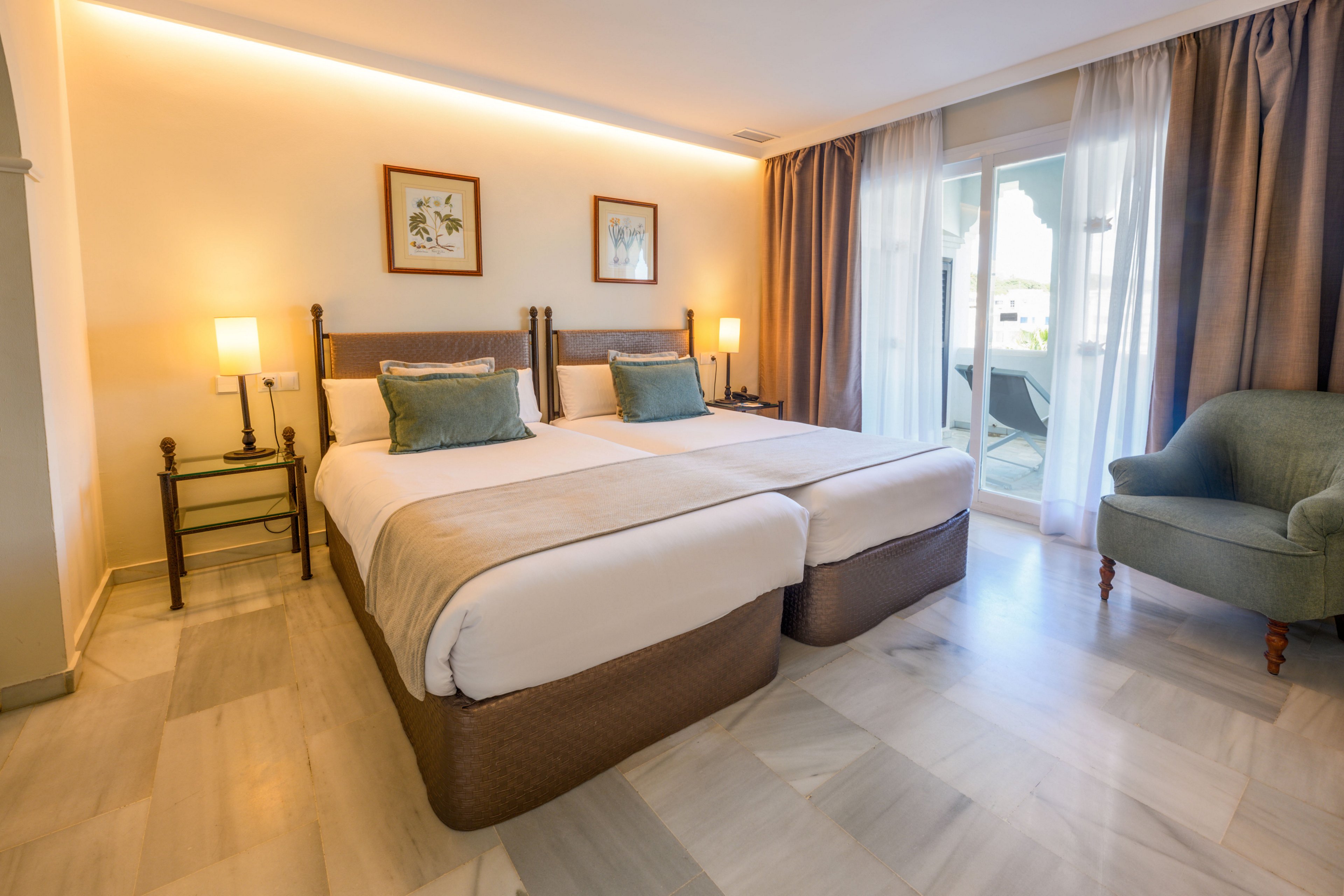 A contemporary hotel room features two neatly made twin beds with white linens and soft green accent pillows. Warm bedside lamps and framed artwork decorate the light-colored walls, creating a cozy atmosphere. Floor-to-ceiling curtains frame a glass door that opens to a balcony or terrace area. A comfortable upholstered armchair sits in the corner on a polished light marble floor, enhancing the clean and inviting look of the space.