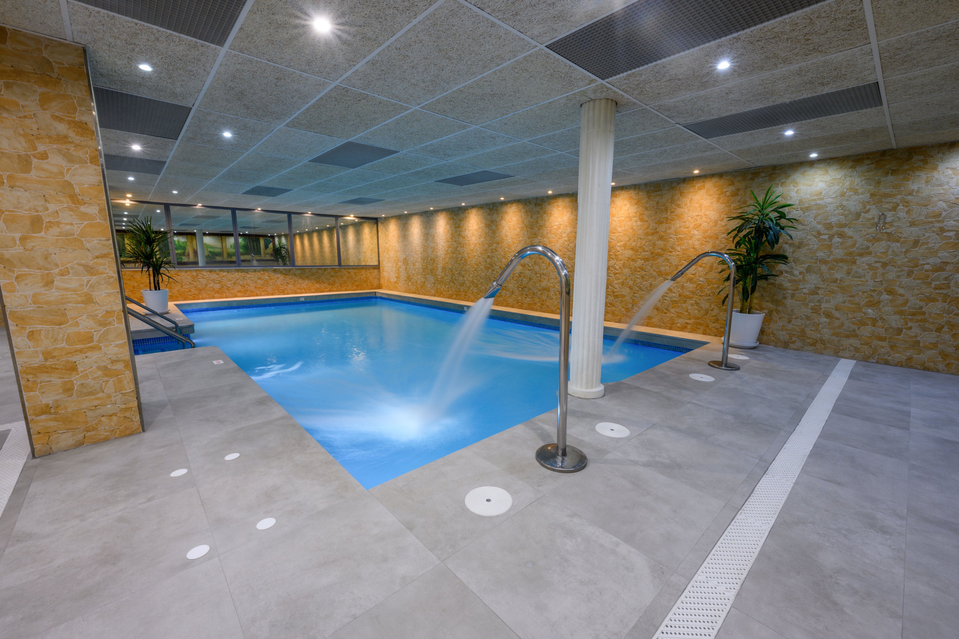 A contemporary indoor swimming pool features powerful stainless steel water jets flowing into clear blue water. The space is surrounded by warm stone-textured walls and a tiled ceiling with recessed lighting, creating a relaxing atmosphere. Large windows line one side of the room, while potted plants add a touch of greenery to the minimalist design. The clean grey floor and white drainage channels emphasize the spa-like, hygienic environment.