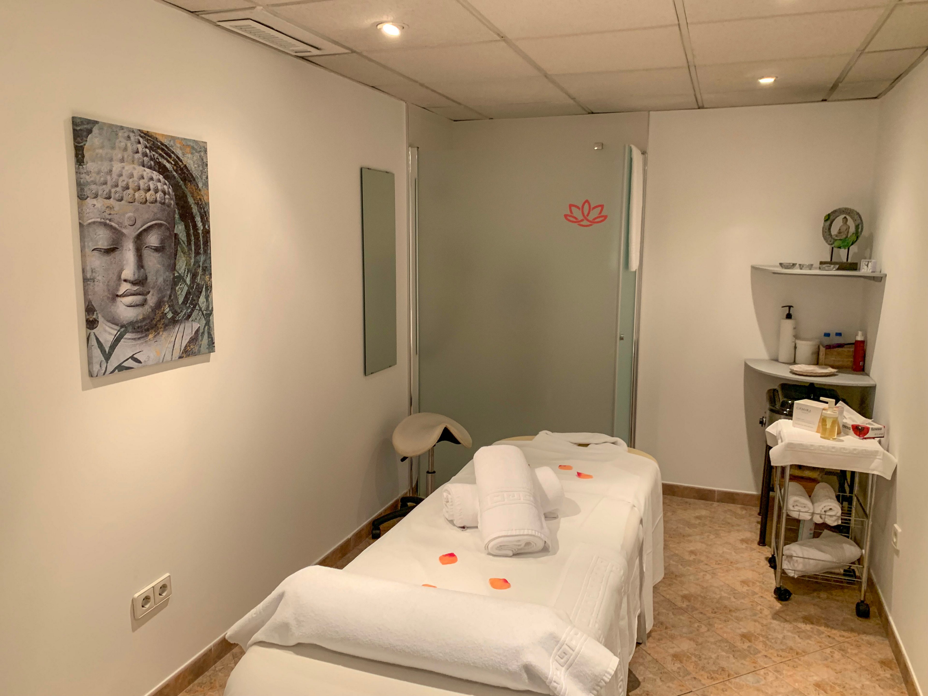 A neatly prepared massage table sits in a small spa treatment room, topped with folded white towels and scattered rose petals. The space features clean white walls, a tiled floor, and soft lighting that creates a tranquil atmosphere. A large Buddha face artwork hangs on the wall, while a frosted glass door with a lotus motif adds to the zen-inspired decor. A side trolley holds spa accessories and supplies, emphasizing a professional wellness setting.