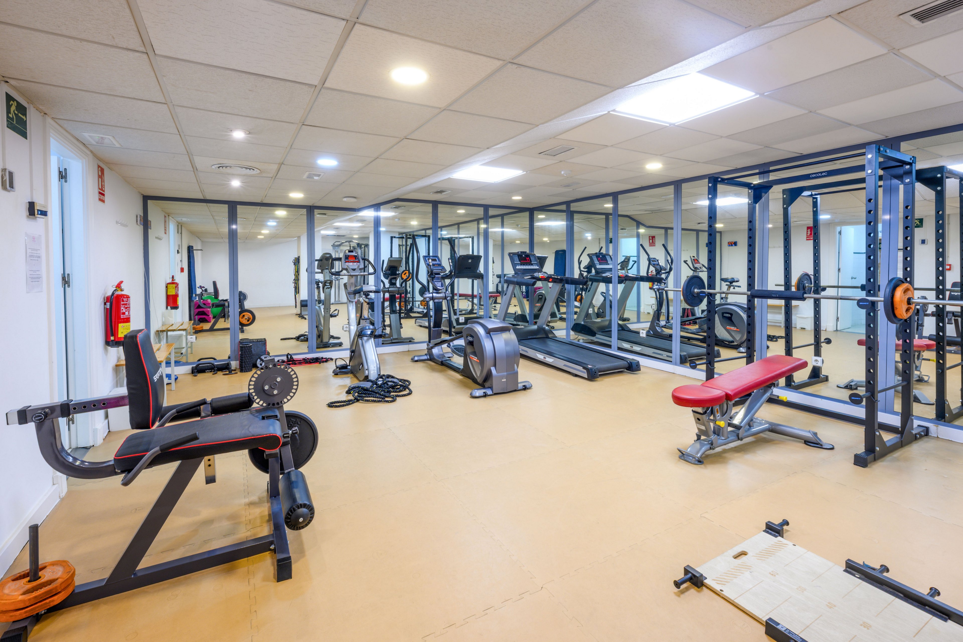 Spacious indoor fitness room featuring a variety of exercise machines and free weights. Cardio equipment such as treadmills and stationary bikes line the mirrored wall, while strength training benches and racks occupy the center. The room has a clean, bright atmosphere with overhead lighting and a neutral floor. No people are present, emphasizing the equipment and organized workout space.