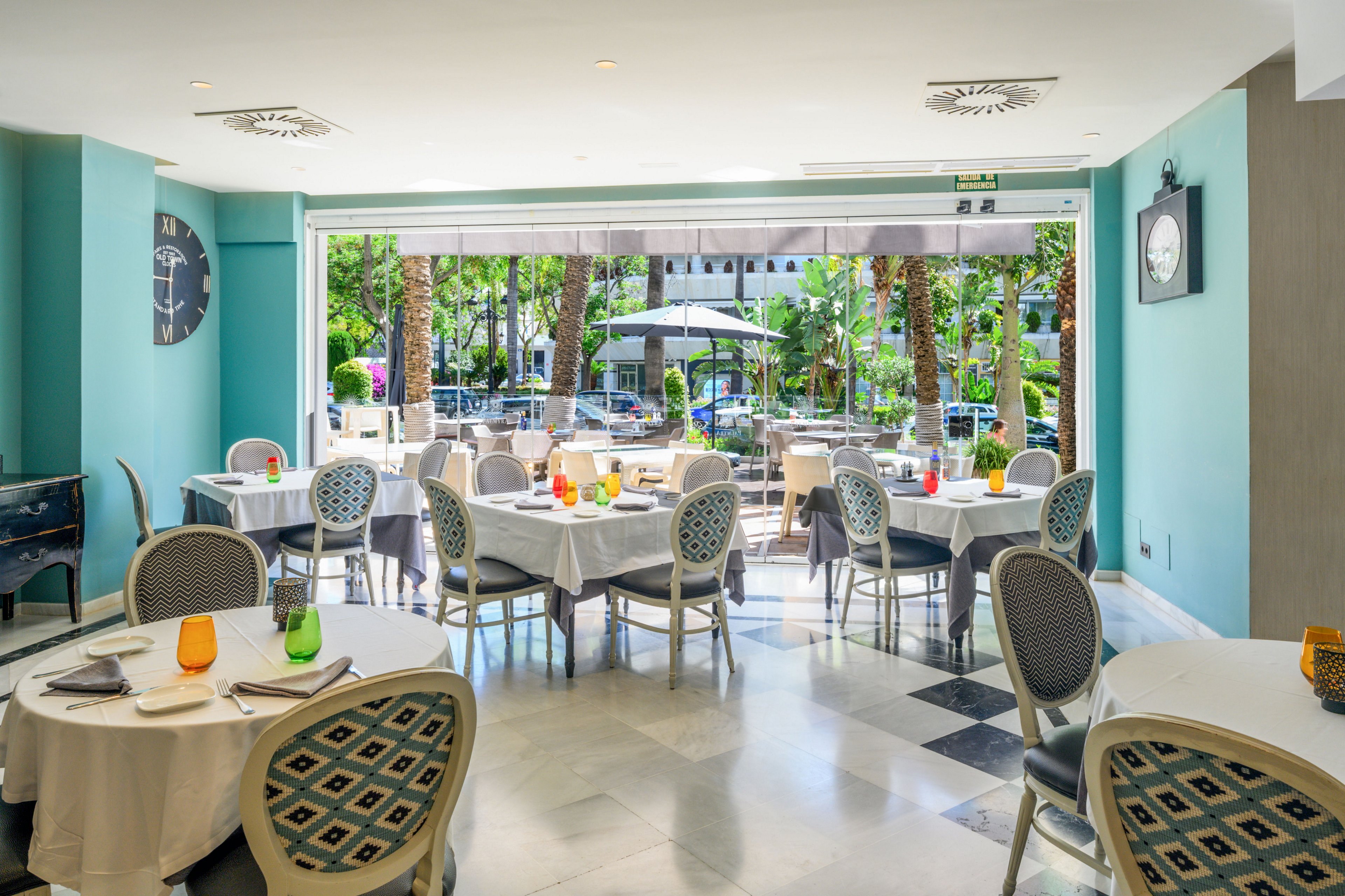 Elegant round tables with white tablecloths are arranged in a bright hotel restaurant interior. Colorful glassware and neatly set place settings add visual interest to the contemporary decor. Large open doors lead to an outdoor terrace with additional tables and lush palm trees, creating a relaxed garden atmosphere. Soft turquoise walls, patterned chairs, and wall clocks contribute to the modern yet inviting dining space.