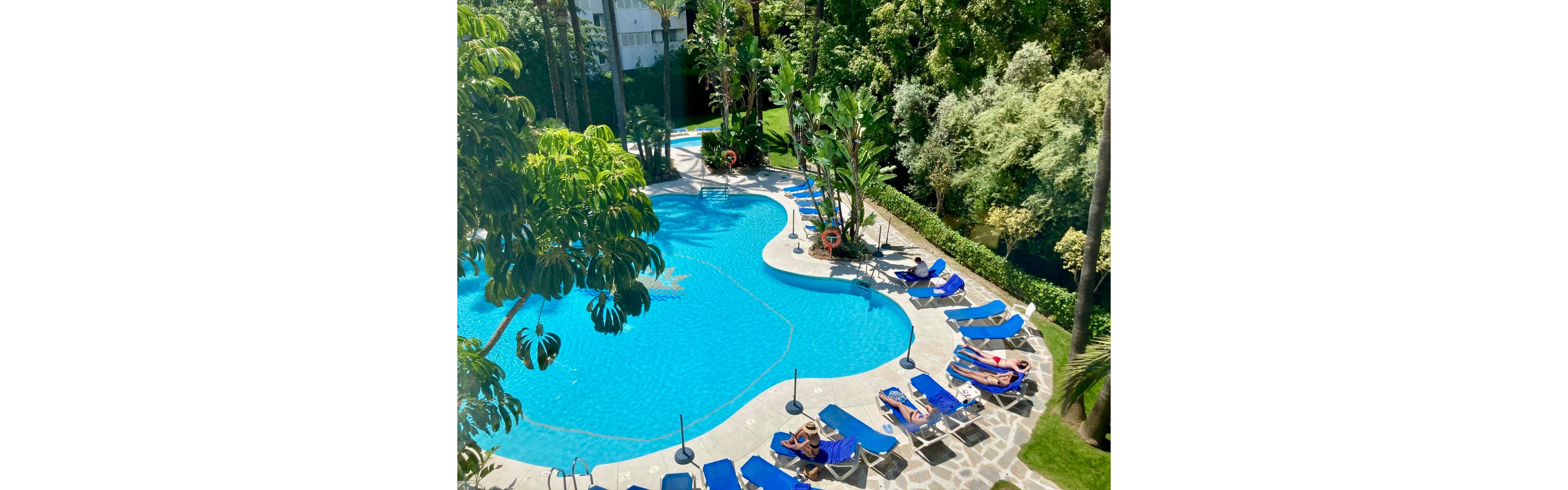 An outdoor resort-style swimming pool is surrounded by blue sun loungers and lush green trees. A few people relax on the loungers and by the poolside in bright sunlight. The pool has a curved, organic shape with clear blue water and several orange life rings on stands. Stone paving and manicured greenery create a tranquil vacation atmosphere.