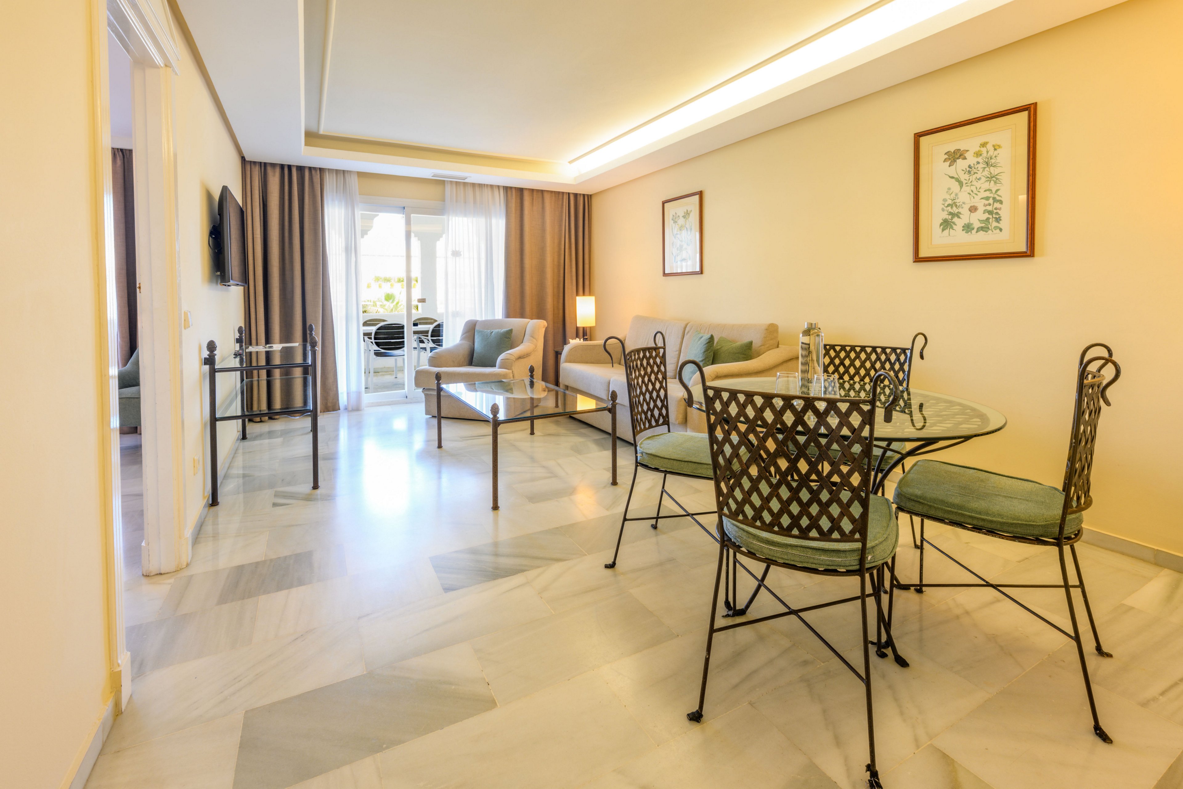 Spacious hotel suite living room with a round dining table and cushioned chairs in the foreground. A comfortable seating area with sofas, coffee table, and floor lamp extends toward large glass doors leading to a balcony. Neutral beige walls, light marble flooring, and soft pastel upholstery create a calm, contemporary atmosphere. Framed artwork and a wall-mounted television add subtle decorative details to the clean interior design.