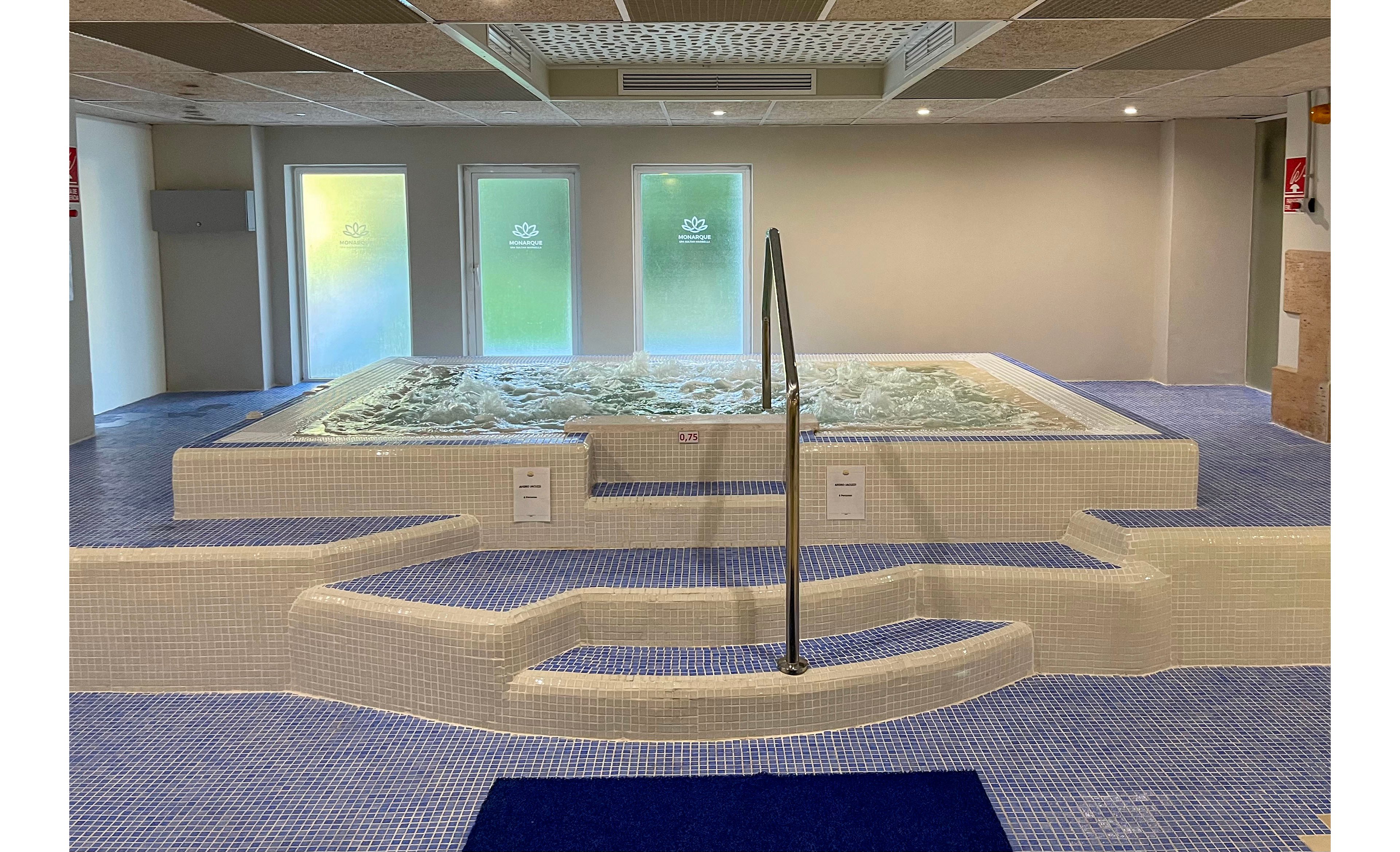 An indoor spa area features a large bubbling jacuzzi pool surrounded by white tiled walls and blue mosaic flooring. A central metal handrail leads down a small set of steps into the water, emphasizing safe access. Frosted glass doors or windows at the back let in soft natural light, creating a calm atmosphere. The clean, modern design and neutral colors highlight a relaxing wellness environment.