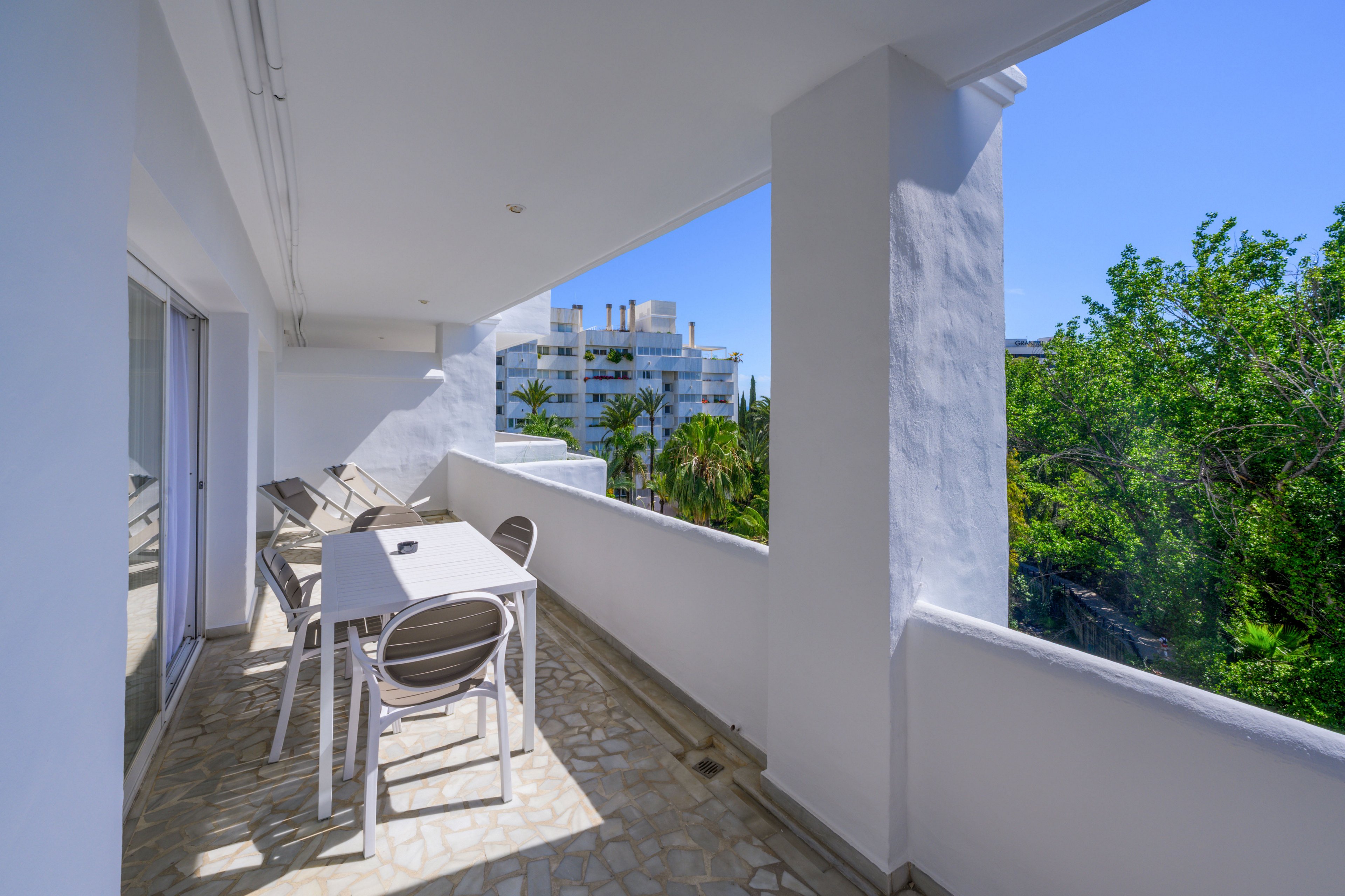 A spacious white balcony features a simple outdoor dining table with several chairs. Sunlight casts strong shadows across the tiled floor, emphasizing the clean, modern architecture. Lush green trees and distant white buildings are visible beyond the balcony wall under a clear blue sky. The scene suggests a relaxing, warm-weather setting ideal for leisure or casual meals outdoors.