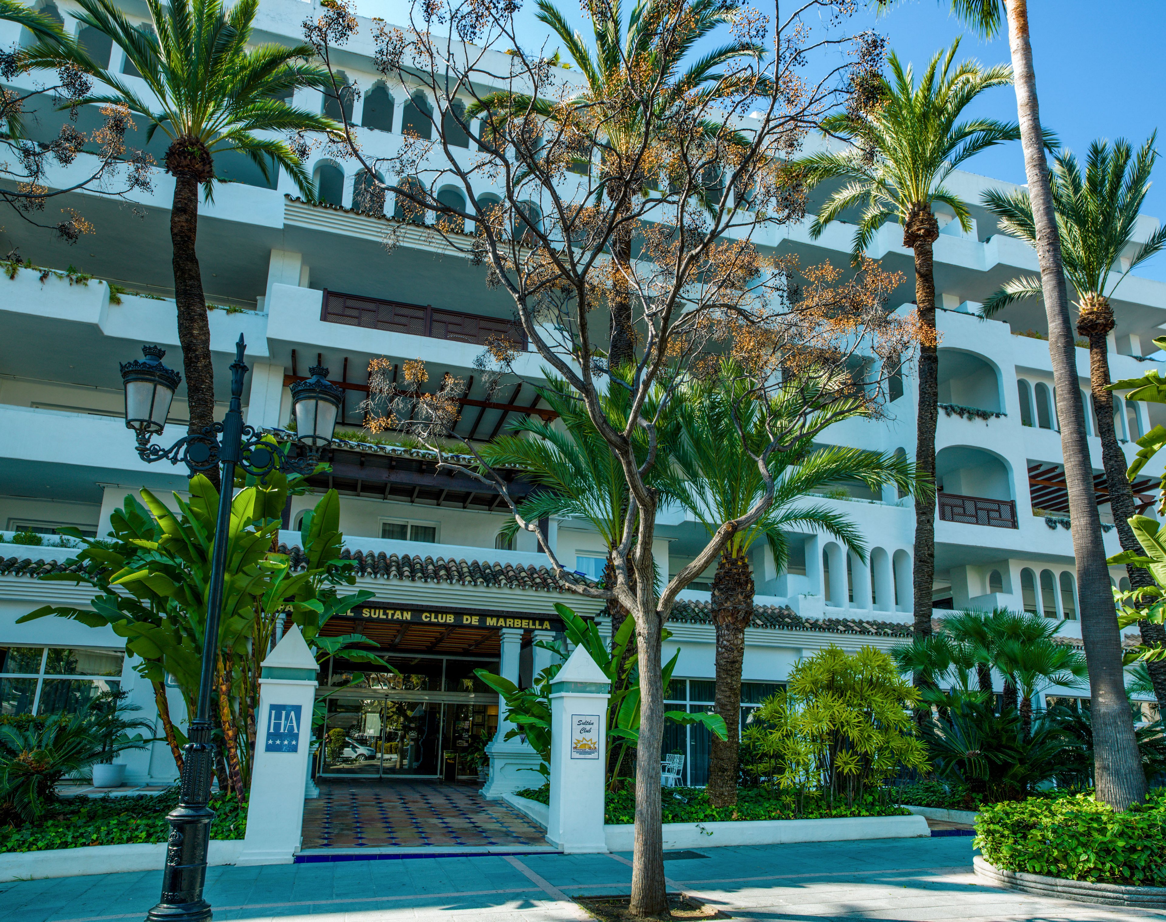 Front view of the Sultan Club de Marbella building surrounded by lush tropical greenery and tall palm trees. The white multi-story structure features balconies, arches, and decorative railings in a Mediterranean architectural style. A tiled entrance walkway leads to glass doors beneath a sign reading "Sultan Club de Marbella." Classic street lamps and manicured plants frame the pathway, creating a welcoming resort atmosphere.
