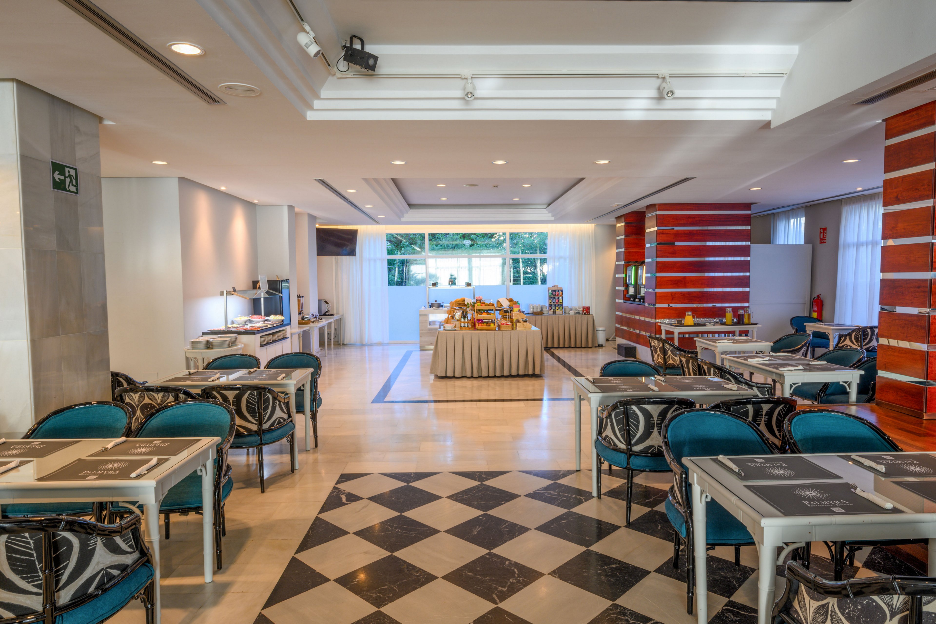 A modern hotel dining room features neatly arranged tables and teal chairs. At the back of the room, a central buffet table is set with breakfast items and drink dispensers. Large windows bring in natural light, illuminating the polished floor and checkered tile pattern in the foreground. The space appears clean, organized, and ready for guests, though no people are present.
