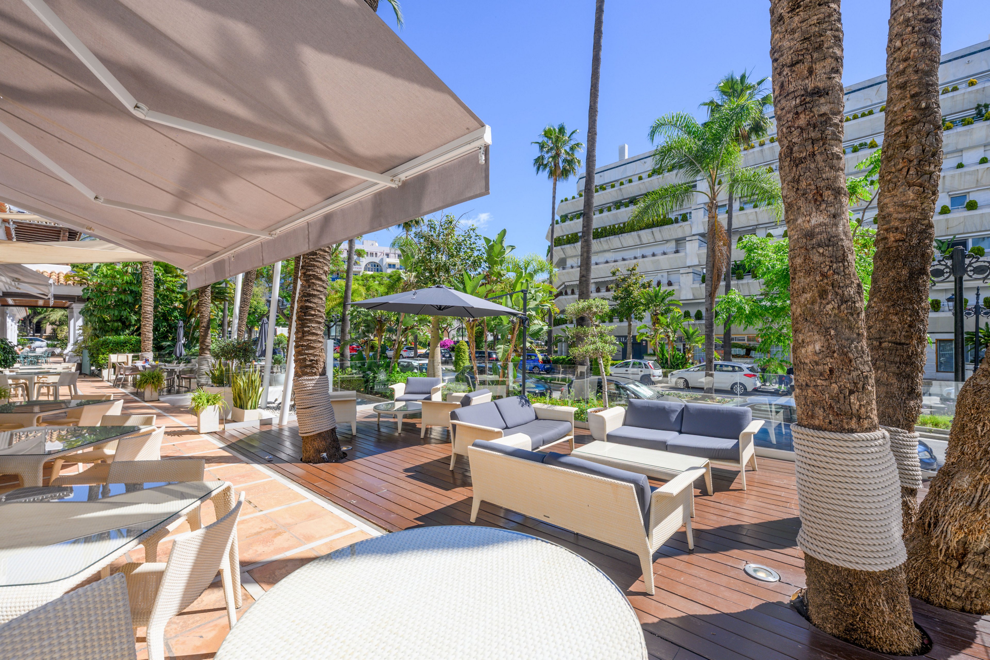 Bright outdoor lounge area with modern white seating and tables is set on a wooden deck under large sunshades. Palm trees and lush greenery surround the terrace, creating a relaxed resort-like atmosphere. Nearby buildings and street elements suggest an urban setting, while the clear blue sky enhances the summery feel. The space appears ready for guests to sit, dine, or enjoy drinks outdoors.