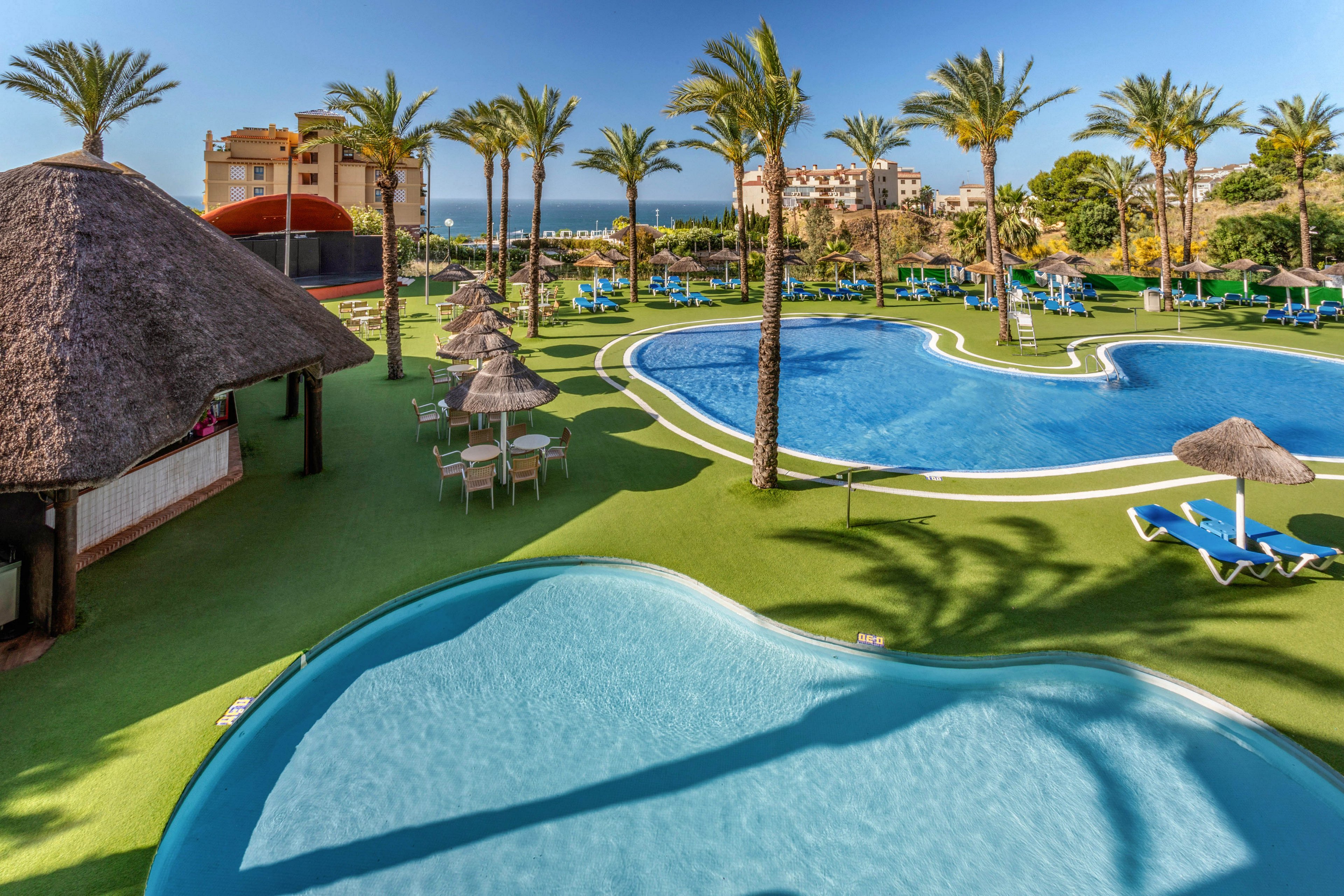 A large resort pool complex features curving blue swimming pools surrounded by bright green artificial grass. Palm trees and thatched umbrellas provide shade over sun loungers arranged neatly around the water. A smaller shallow pool sits in the foreground, while apartment-style buildings and the sea are visible in the distance. The clear sky and vibrant colors create a warm, relaxing vacation atmosphere.