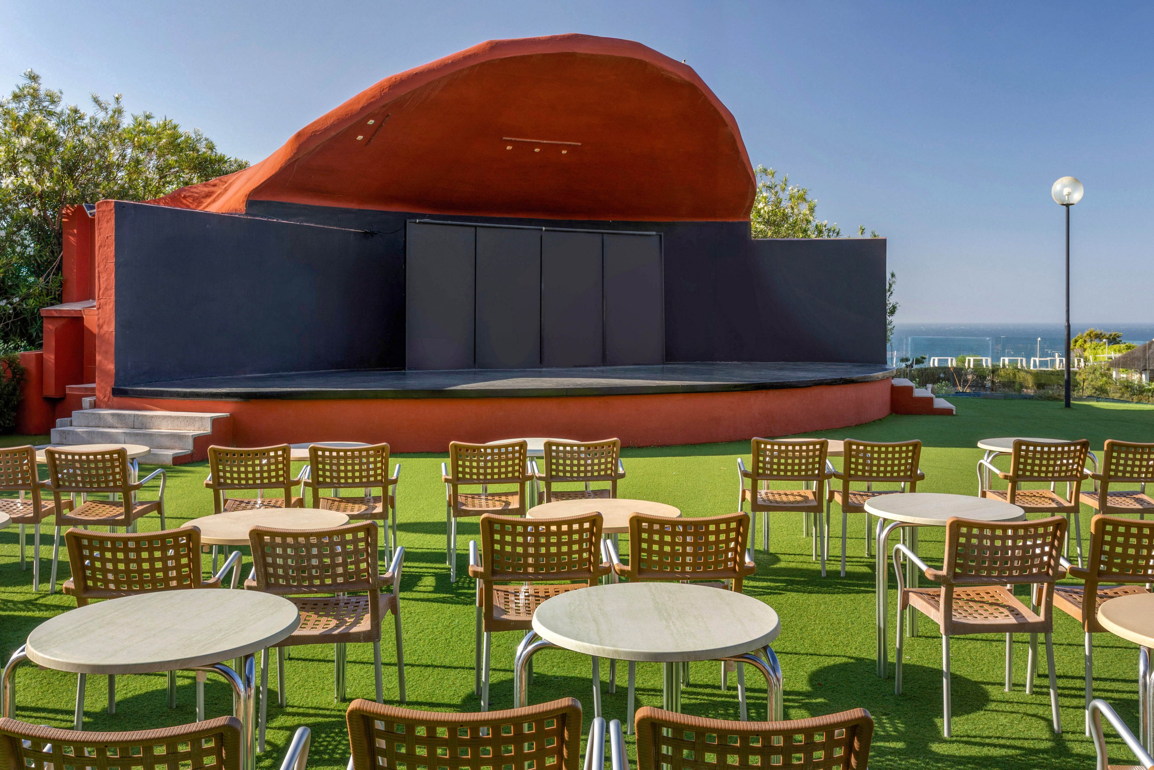 An outdoor performance stage with a curved red backdrop stands at the center of a sunny open-air venue. Rows of wicker-style chairs and round metal tables are arranged on a neatly maintained green lawn facing the stage. Trees and shrubs frame the area, and a glimpse of the sea and clear blue sky is visible in the background. The scene is calm and quiet, suggesting preparation for an upcoming show or event.
