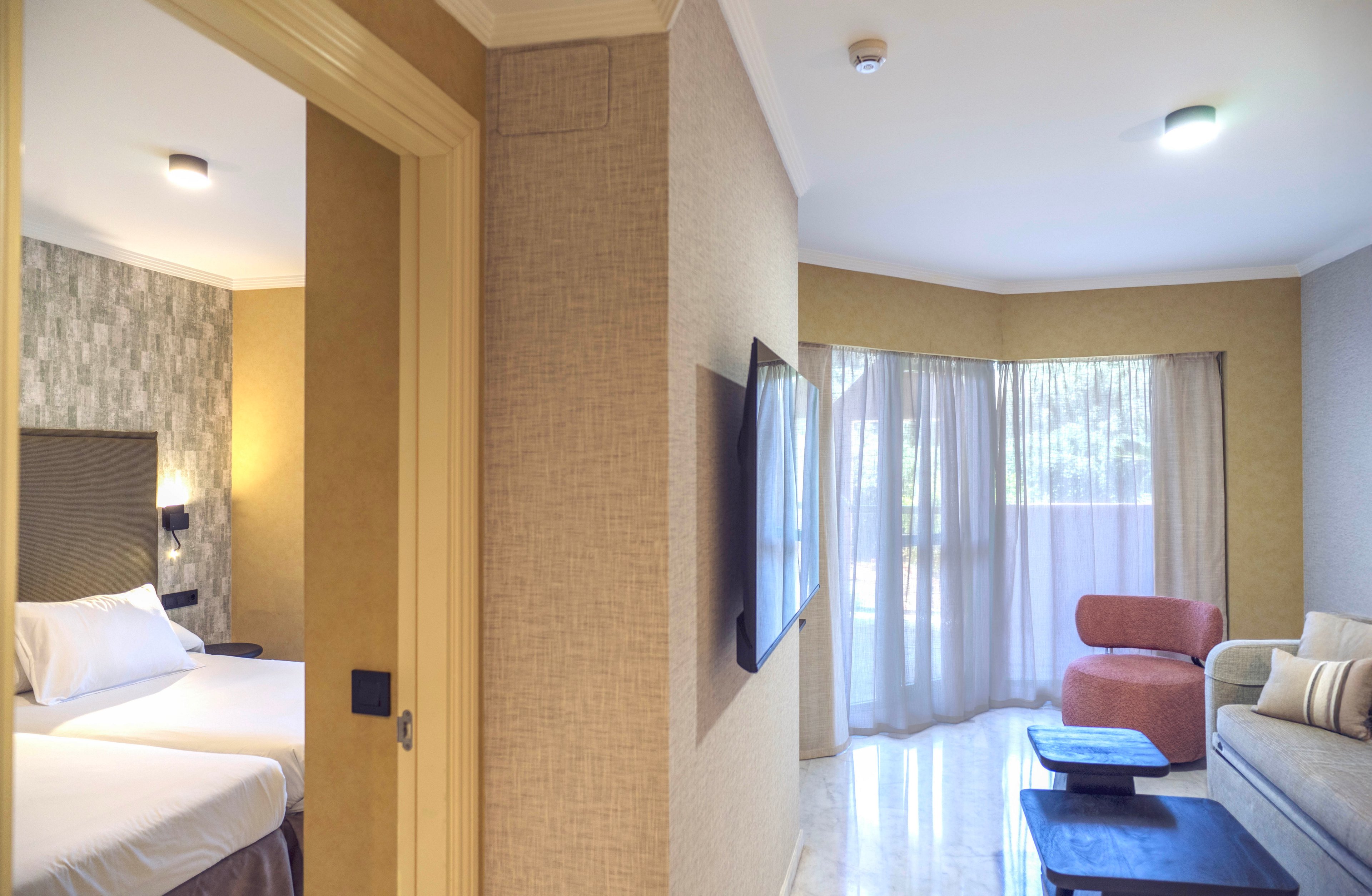 The image shows a contemporary hotel suite featuring a cozy bedroom with white linens and a stylish headboard. Adjacent to the bedroom, the lounge area includes a sofa, armchair, coffee tables, and a wall-mounted television. Large windows with sheer curtains allow natural light to fill the space, highlighting the neutral color palette and modern decor. The setting appears clean, inviting, and comfortable.