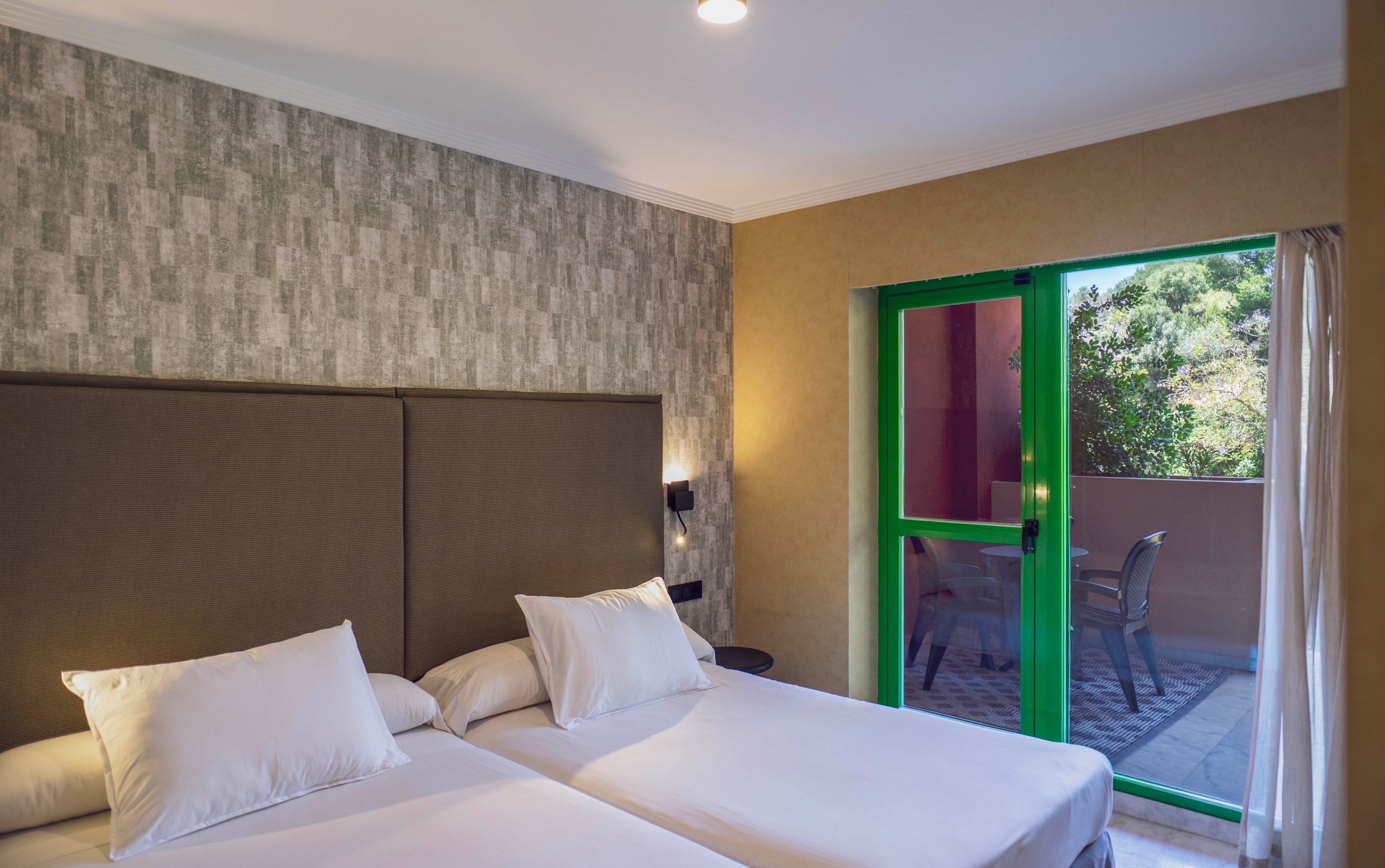 A contemporary hotel room features two twin beds with white linens and a large padded headboard. The room has a textured accent wall and a green-framed glass door leading to a balcony with outdoor seating. Natural light fills the space, highlighting the neutral color palette and minimalist decor.
