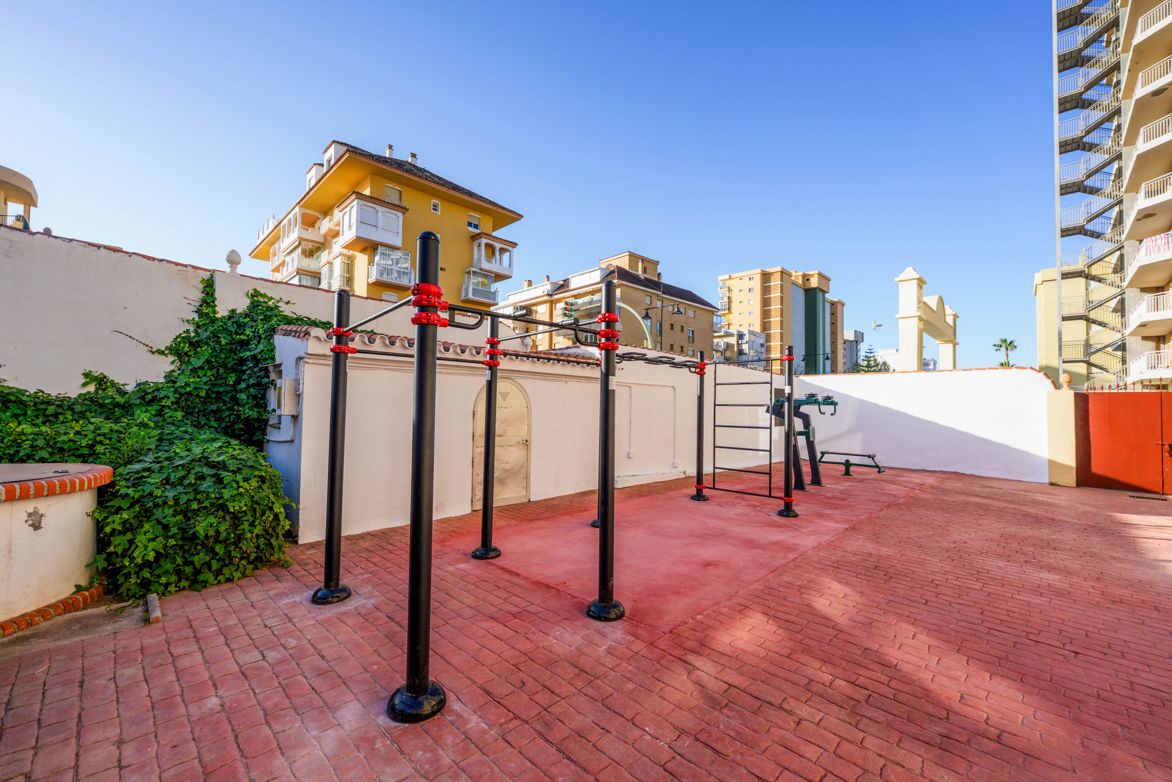 An outdoor calisthenics workout area is set up on a red brick courtyard surrounded by white walls. Several metal pull-up bars and parallel bars stand in the middle of the space, with additional exercise equipment visible in the background. Residential and hotel buildings rise around the courtyard under a clear blue sky. Green ivy climbs one of the walls, adding a touch of nature to the urban fitness setting.