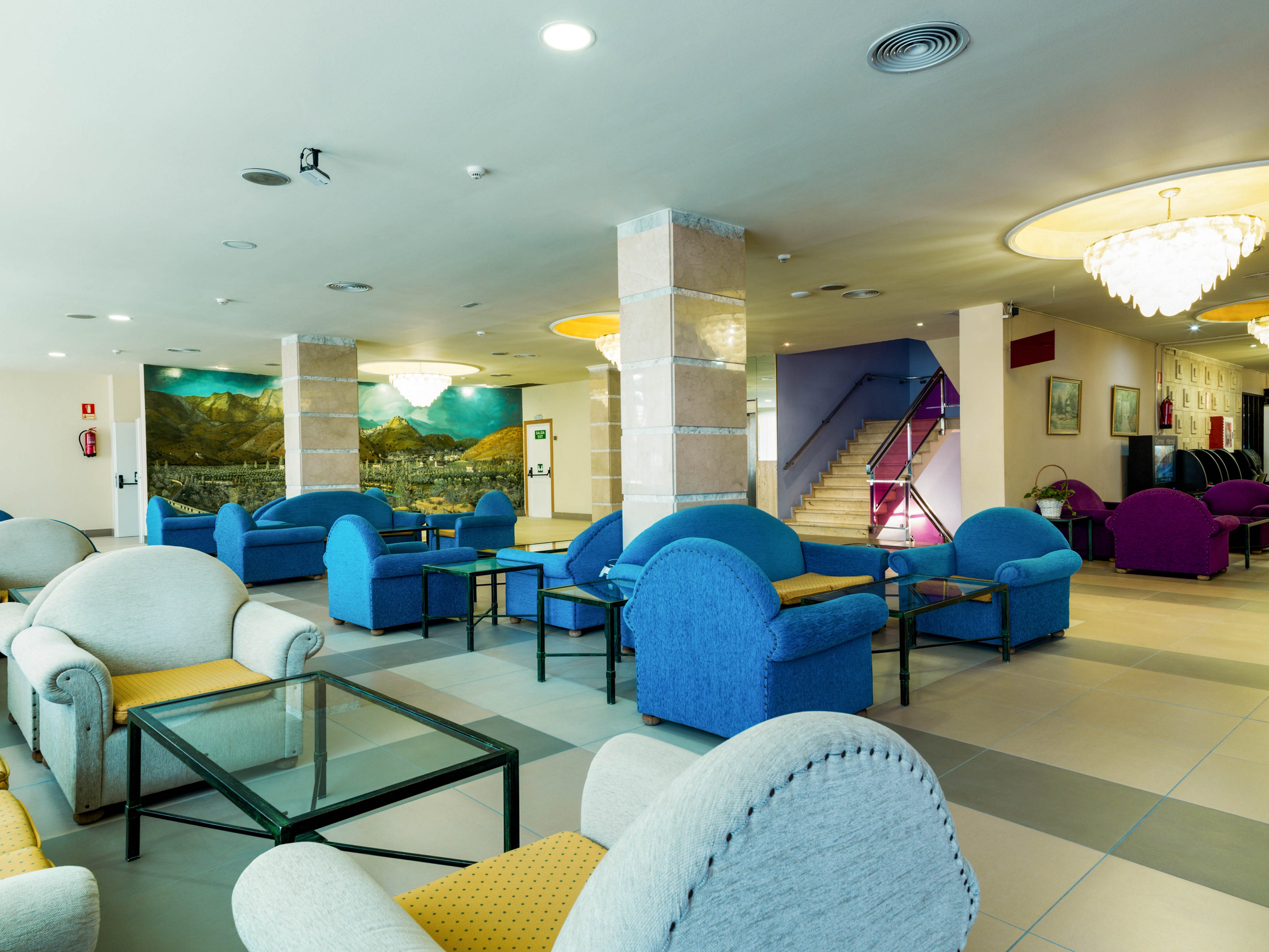 A spacious hotel lobby features an array of colorful armchairs and sofas arranged around glass coffee tables. The setting includes tiled floors, large supporting columns, and a mural of a mountainous landscape on the back wall. A staircase with a glass railing leads to an upper level, while chandeliers and recessed lighting brighten the space. The overall atmosphere is modern, welcoming, and comfortable for guests.