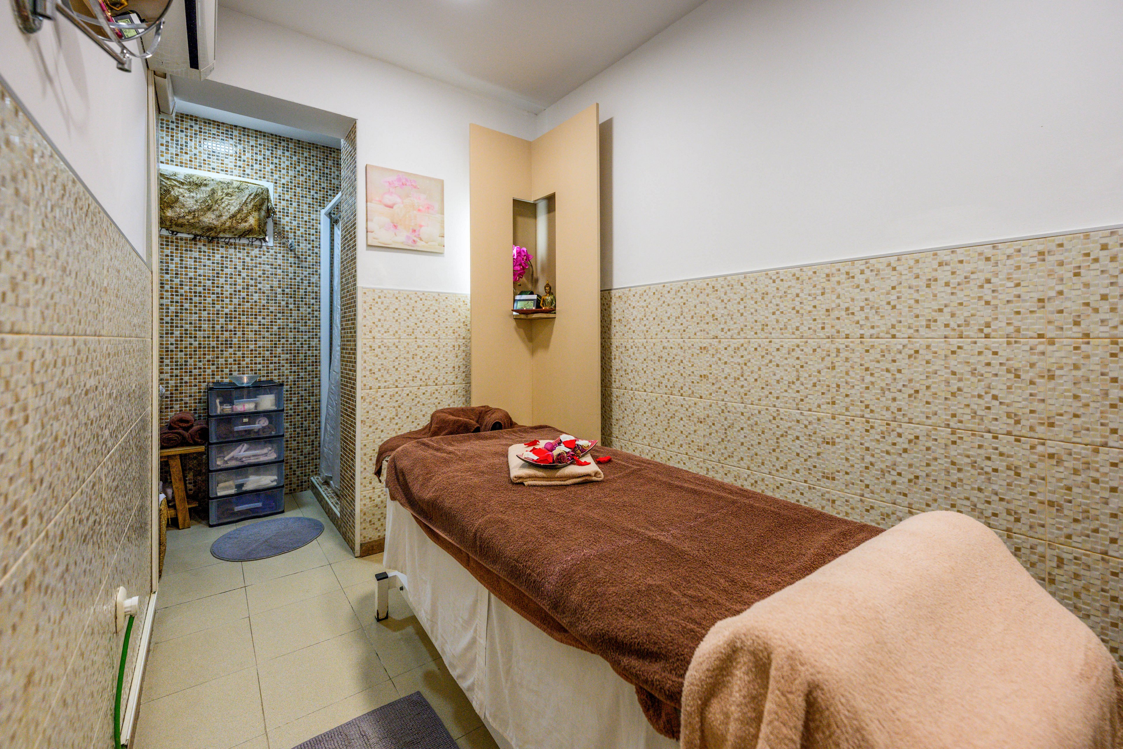 A small spa treatment room features a massage table covered with a brown blanket and neatly folded towels. The narrow space is lined with patterned wall tiles and soft lighting, creating a warm and inviting atmosphere. A shelving niche with decorative items and a plastic drawer unit in the back add functional storage. The overall setting suggests a relaxing environment for massage or wellness treatments.