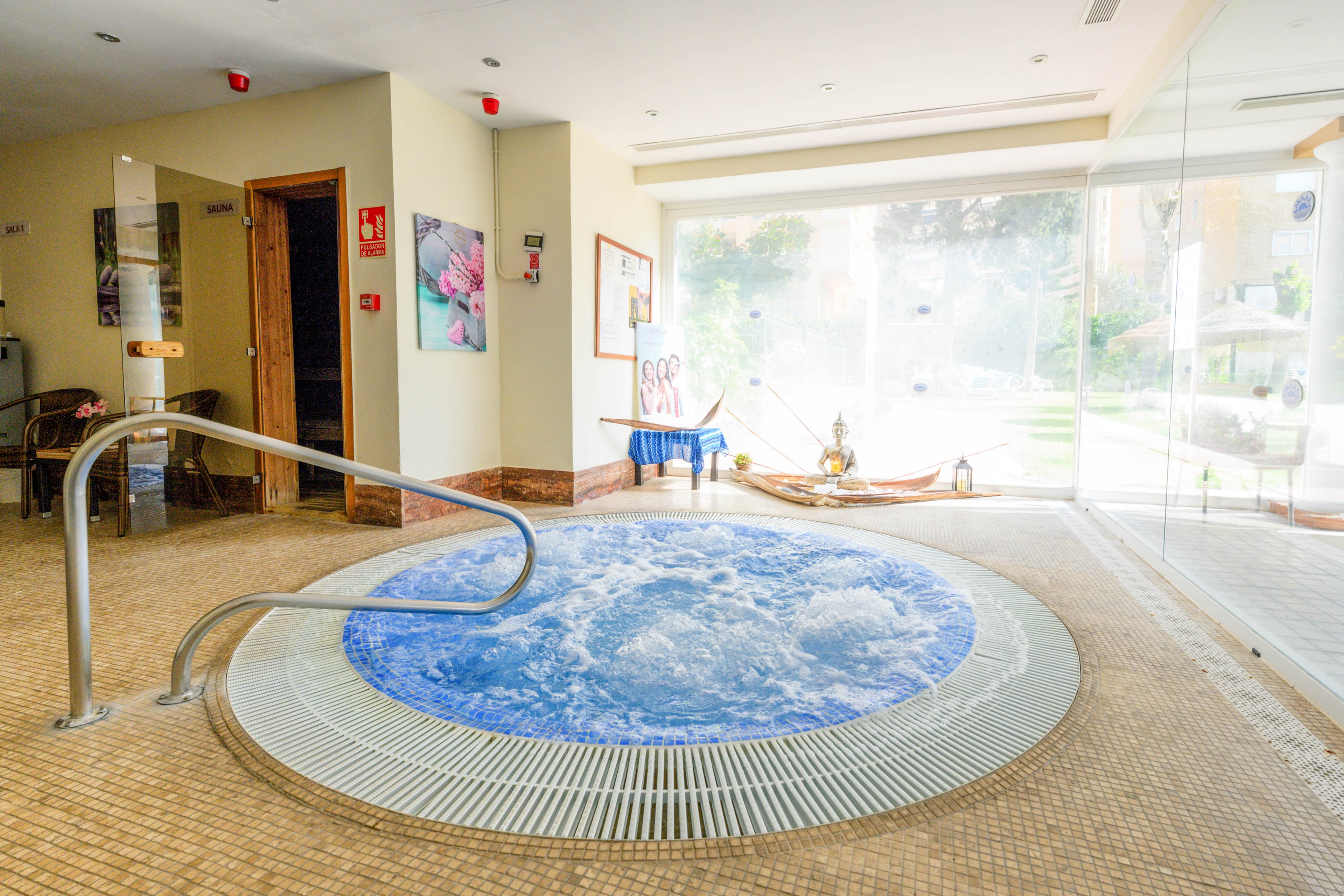 An indoor circular jacuzzi filled with bubbling water is the main focus of this spa room. The hot tub is surrounded by a tiled floor and features a metal handrail for safe entry. Large floor-to-ceiling windows reveal a bright garden area outside, allowing natural light to flood the space. Wall art, safety signs, and spa decor contribute to a calm, wellness-focused atmosphere.