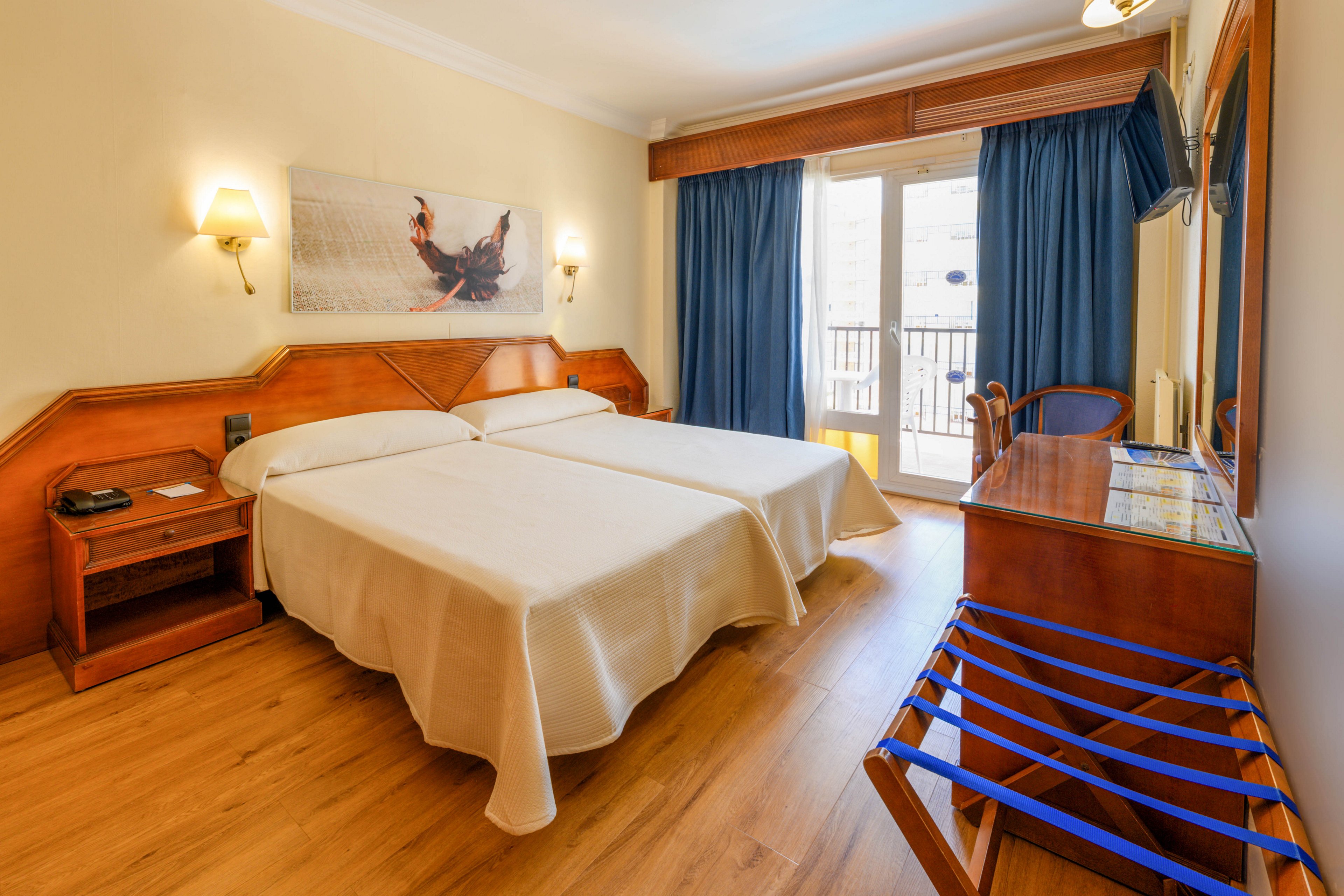 A tidy hotel room features two neatly made twin beds with light-colored bedspreads. Warm wooden furniture, including bedside tables and a dresser with mirror, complements the hardwood floor. Blue curtains frame a glass door leading to a balcony, allowing natural light to fill the space. Wall-mounted lamps and a framed artwork above the beds add a cozy, welcoming atmosphere.