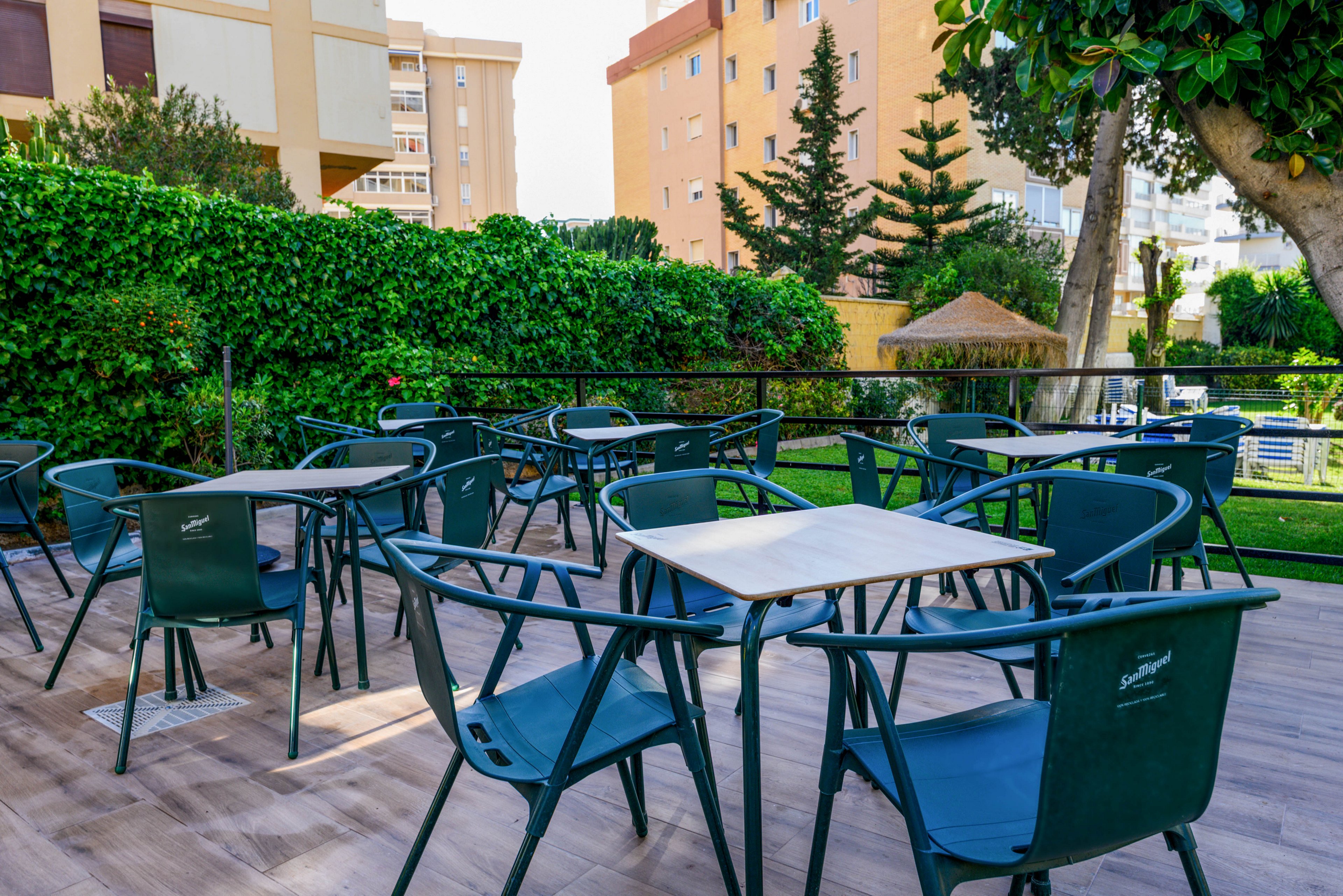 An empty outdoor cafe terrace features several dark green plastic chairs and square tables arranged on a wooden deck. The seating area is bordered by a lush green hedge and trees, creating a cozy garden atmosphere. In the background, multi-story residential or hotel buildings rise above the foliage. Bright daylight and clear weather give the scene a relaxed, inviting feel.