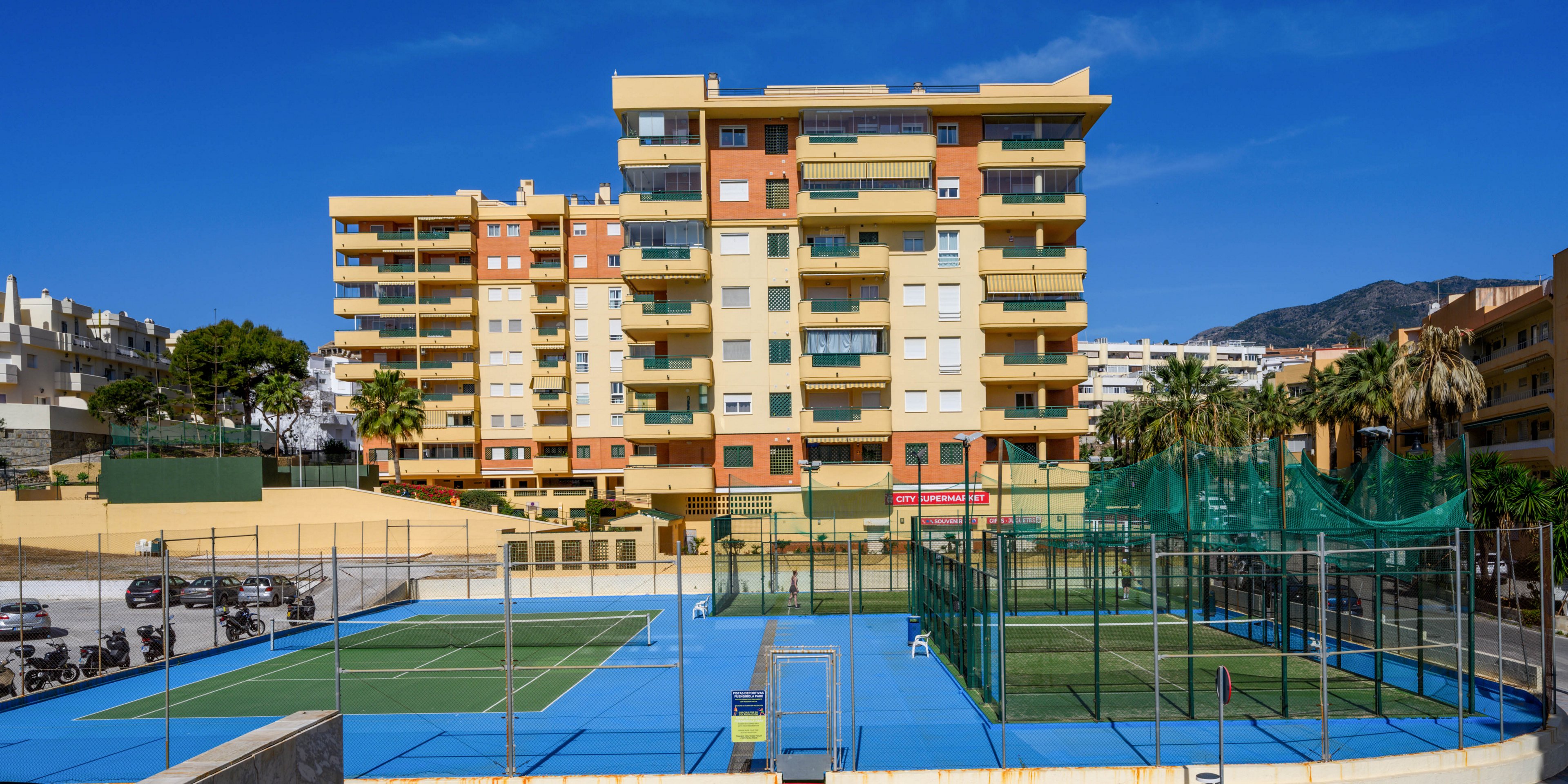 A row of mid-rise residential apartment buildings stands behind a complex of bright blue tennis and sports courts. The scene appears to be in a sunny urban neighborhood with palm trees and a clear blue sky. Multiple fenced courts and nets dominate the foreground, while scooters or motorbikes are parked along the side. Distant hills or mountains rise in the background, adding depth to the cityscape.