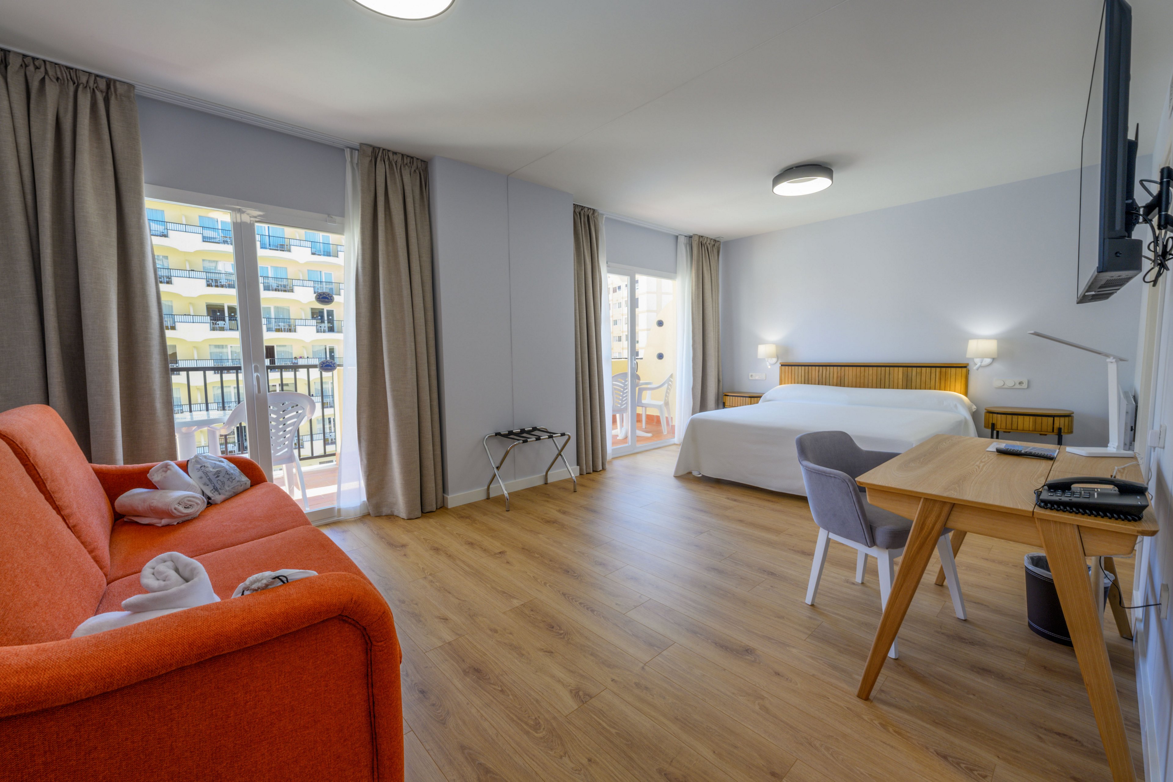 Spacious contemporary hotel room featuring a large bed, wooden flooring, and a work desk with chair. An orange sofa with neatly folded towels adds a pop of color to the neutral decor. Floor-to-ceiling windows with curtains open to a balcony overlooking neighboring apartment buildings. The room is well lit with ceiling fixtures and bedside lamps, creating a clean and inviting atmosphere.
