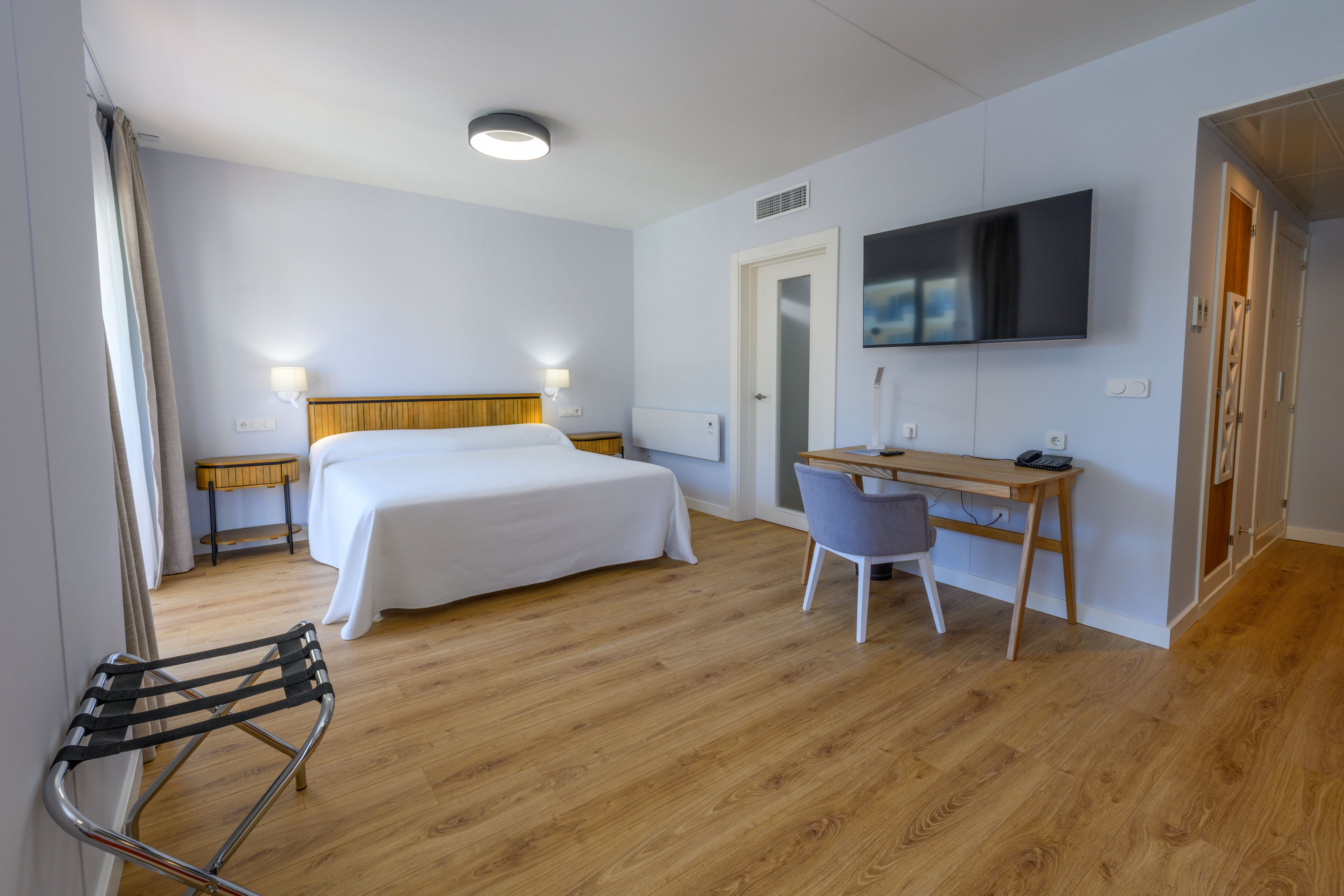 A spacious hotel room features a large bed with white linens against a light-colored wall. Wooden bedside tables and a matching desk with a chair add warmth to the minimalist decor. A flat-screen TV is mounted on the wall opposite the bed, and a luggage rack stands near the window with long curtains. The room has clean lines, ample floor space, and a calm, contemporary atmosphere.
