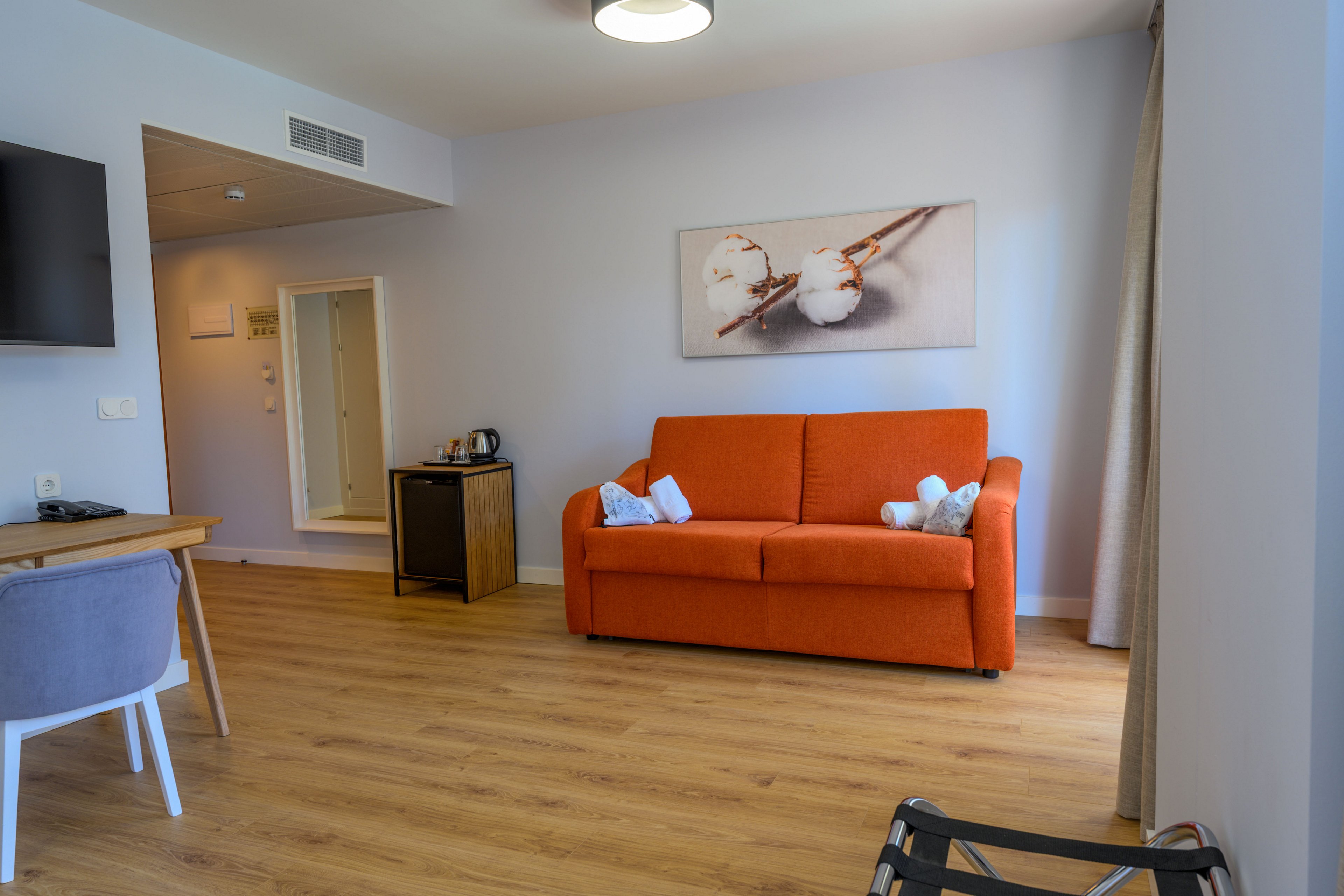 A contemporary hotel room features a bright orange sofa against a light-colored wall. Above the sofa hangs a framed artwork depicting cotton flowers, adding a soft decorative touch. The space includes a wooden floor, a small desk with a chair, and a side table with a kettle and cups. Neutral walls, simple furnishings, and natural light from a window create a clean, welcoming atmosphere.