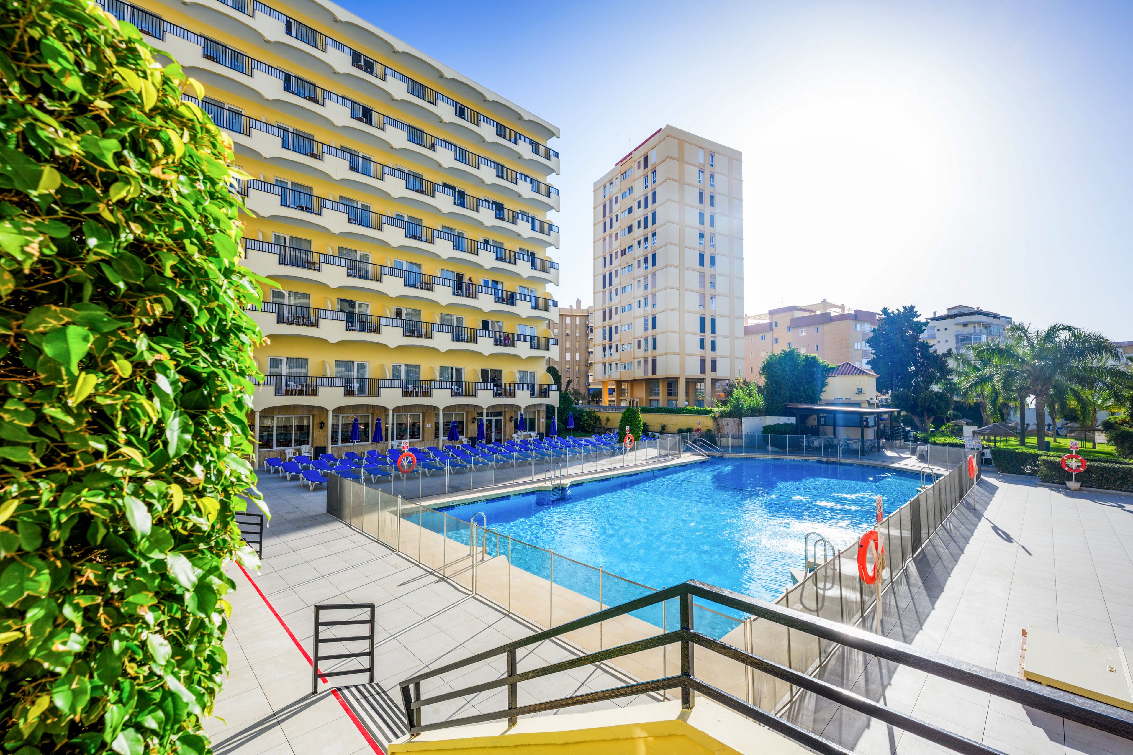 A bright hotel courtyard features a large outdoor swimming pool surrounded by a safety fence. Multi-story residential and hotel buildings frame the scene under a clear blue sky. Blue lounge chairs line the pool deck, and a hedge and palm trees add greenery to the modern urban setting. The overall atmosphere is sunny, relaxed, and inviting for vacation guests.