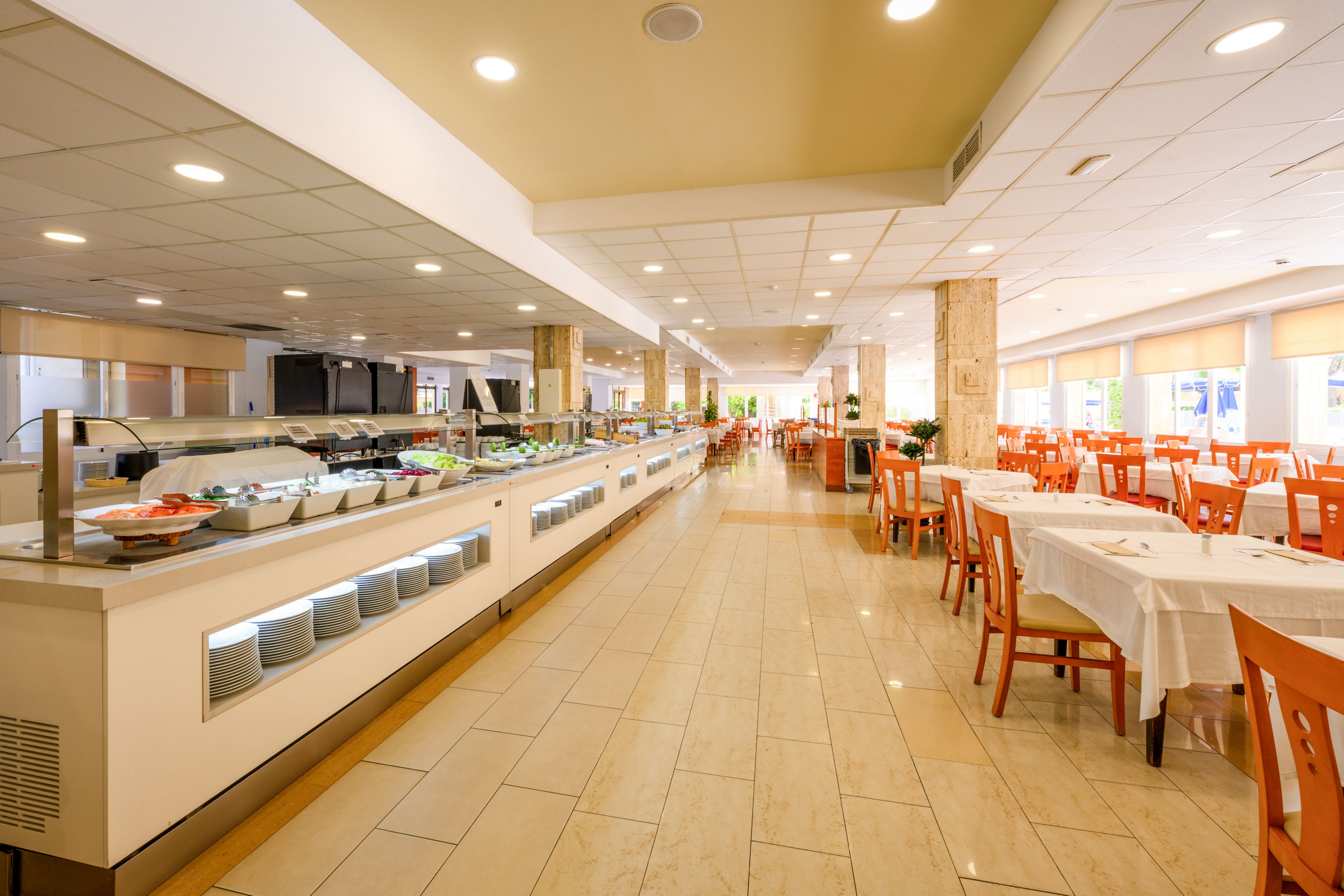 Wide view of a spacious self-service buffet area in a modern restaurant. A long counter displays various dishes and salads in trays under glass. The dining room is filled with neatly arranged tables and orange chairs, all currently unoccupied. Warm lighting, tiled floors, and large windows create an inviting and clean atmosphere.