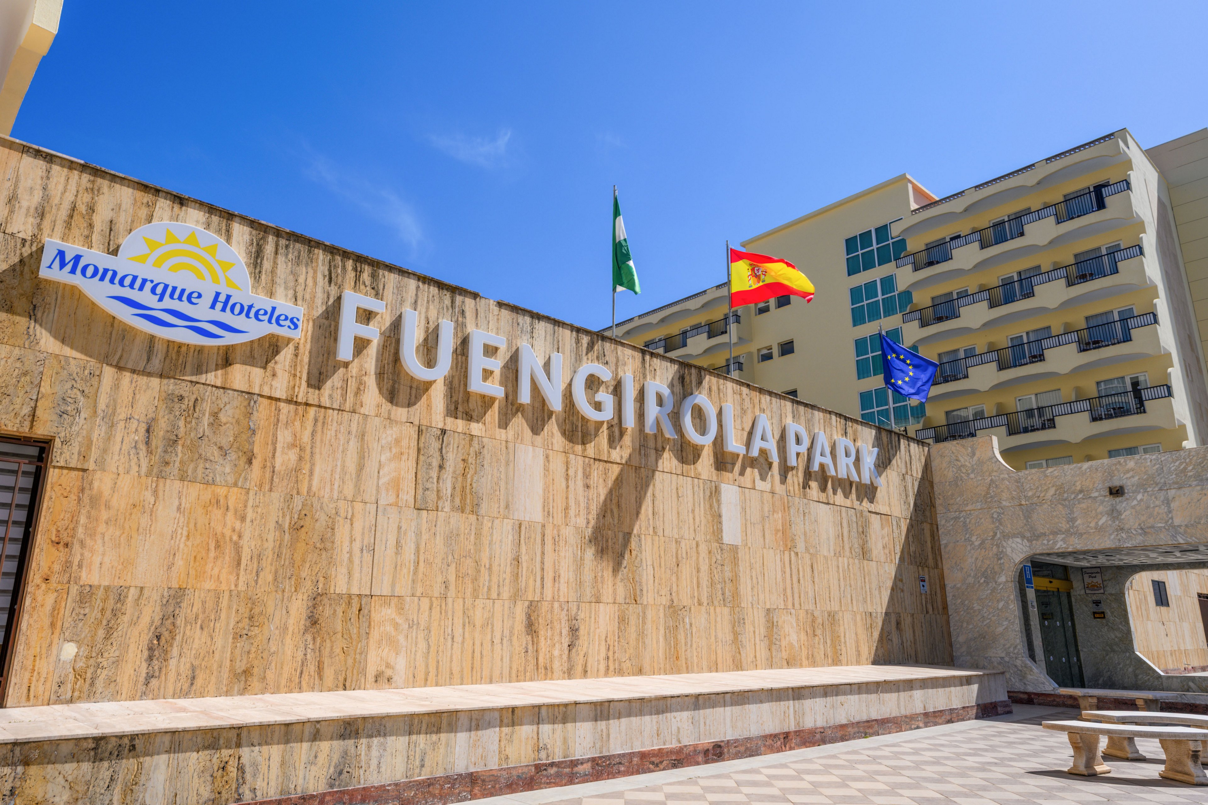Large stone wall signage reading FUENGIROLA PARK marks the entrance to a hotel complex under a clear blue sky. The Monarque Hoteles sign is visible on the left side of the wall. Three flags, including the Spanish flag and the European Union flag, fly above the structure. A multi-story building with balconies rises in the background, suggesting a sunny coastal vacation destination.