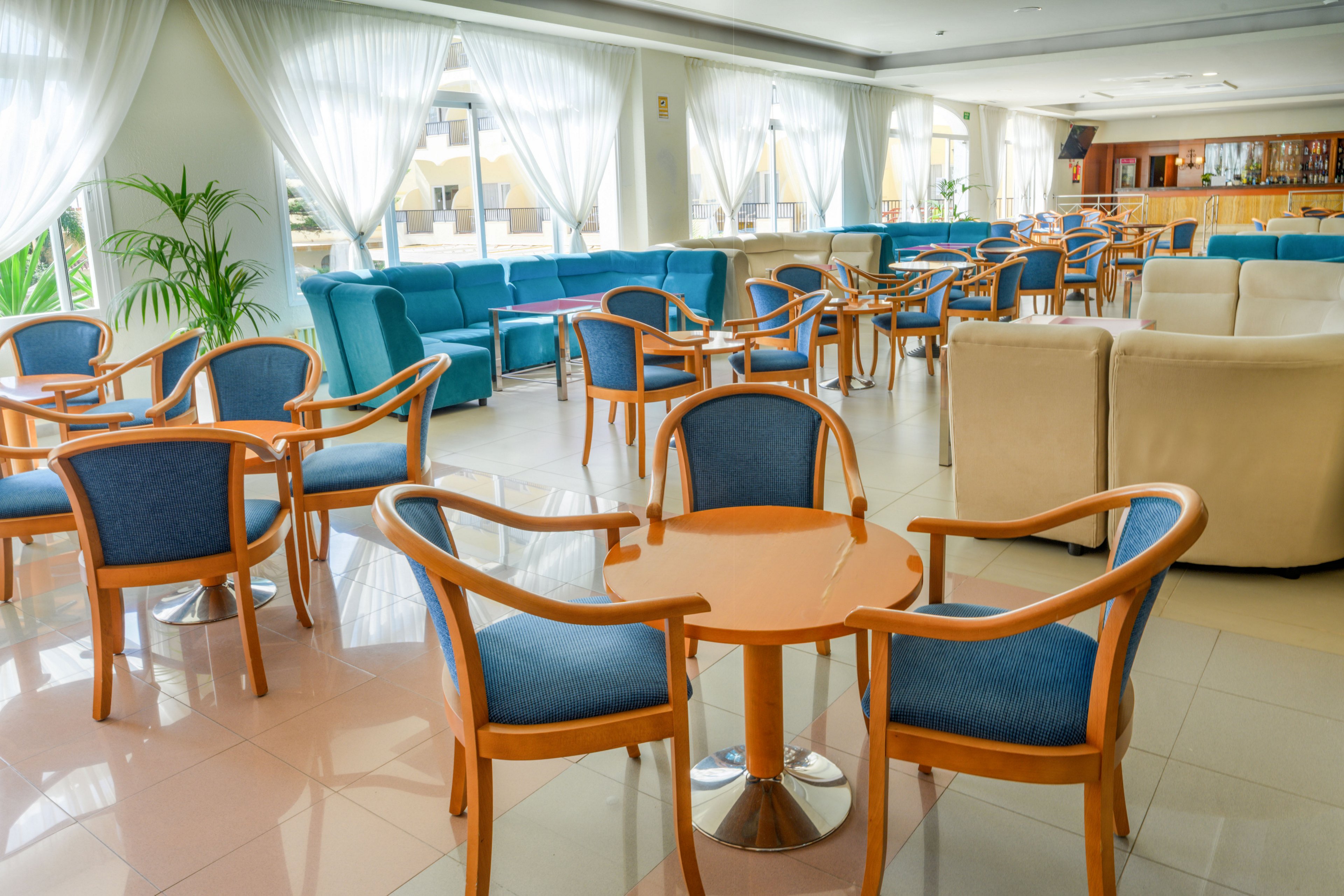 Spacious hotel lounge area with wooden tables and blue upholstered chairs arranged neatly across a tiled floor. Large windows with sheer white curtains allow abundant natural light to fill the room. In the background, comfortable beige and turquoise sofas create relaxed seating zones near a bar counter. The overall atmosphere is clean, airy, and welcoming, suitable for socializing or casual meetings.