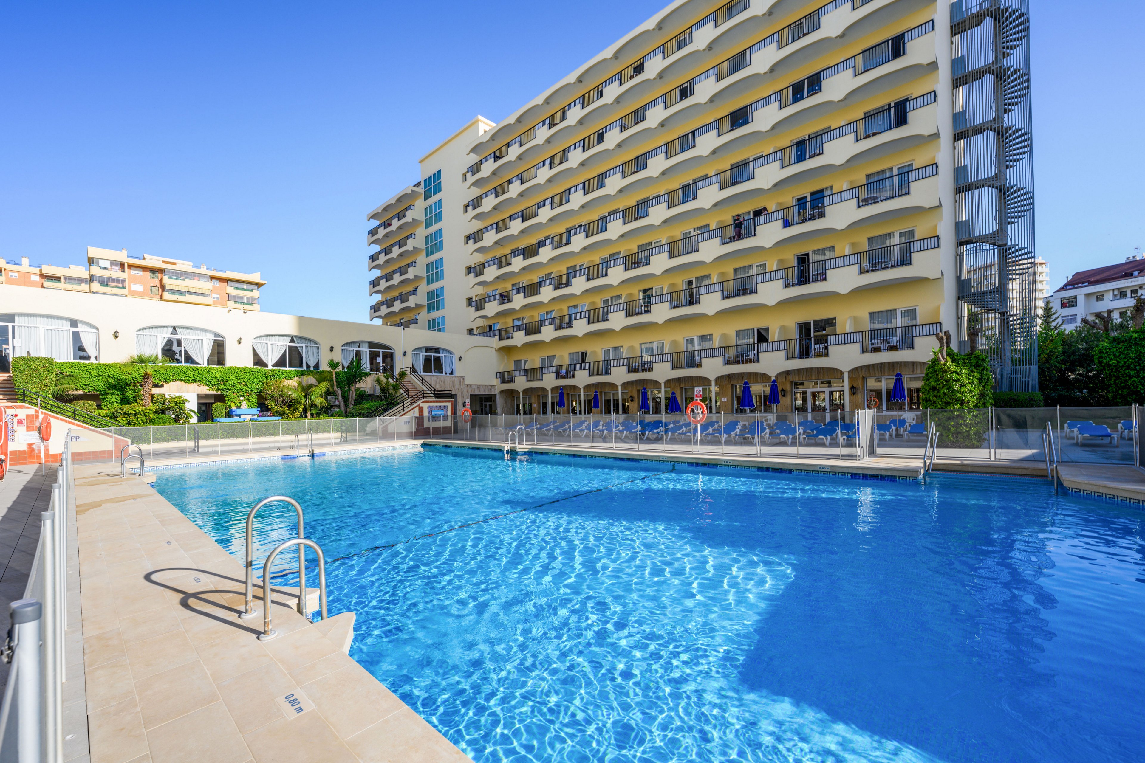 A large outdoor swimming pool sits in front of a multi-story hotel building under a clear blue sky. Sun loungers and umbrellas line the poolside area, suggesting a relaxing vacation setting. The hotel facade features repeating balconies and windows, with greenery and fencing surrounding the pool. Nearby buildings are visible in the background, indicating an urban or resort neighborhood.
