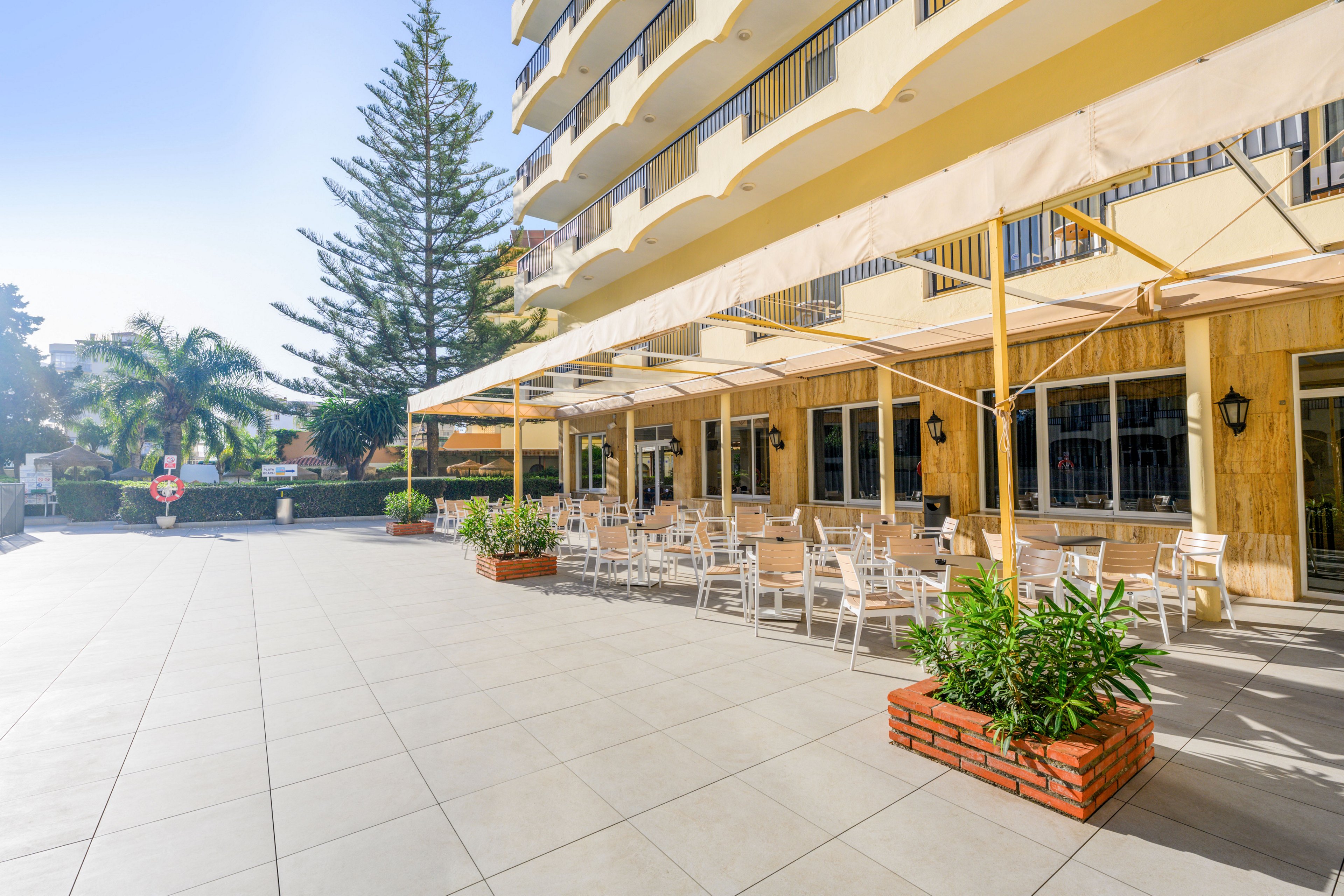 A spacious hotel terrace features numerous white tables and chairs arranged under large beige awnings. The setting is bright and sunny, with clean tiled flooring extending across the open courtyard. Potted plants in brick planters add greenery, while palm trees and other foliage line the background. The building’s balconies and large windows frame the inviting outdoor dining area, suggesting a relaxed vacation atmosphere.