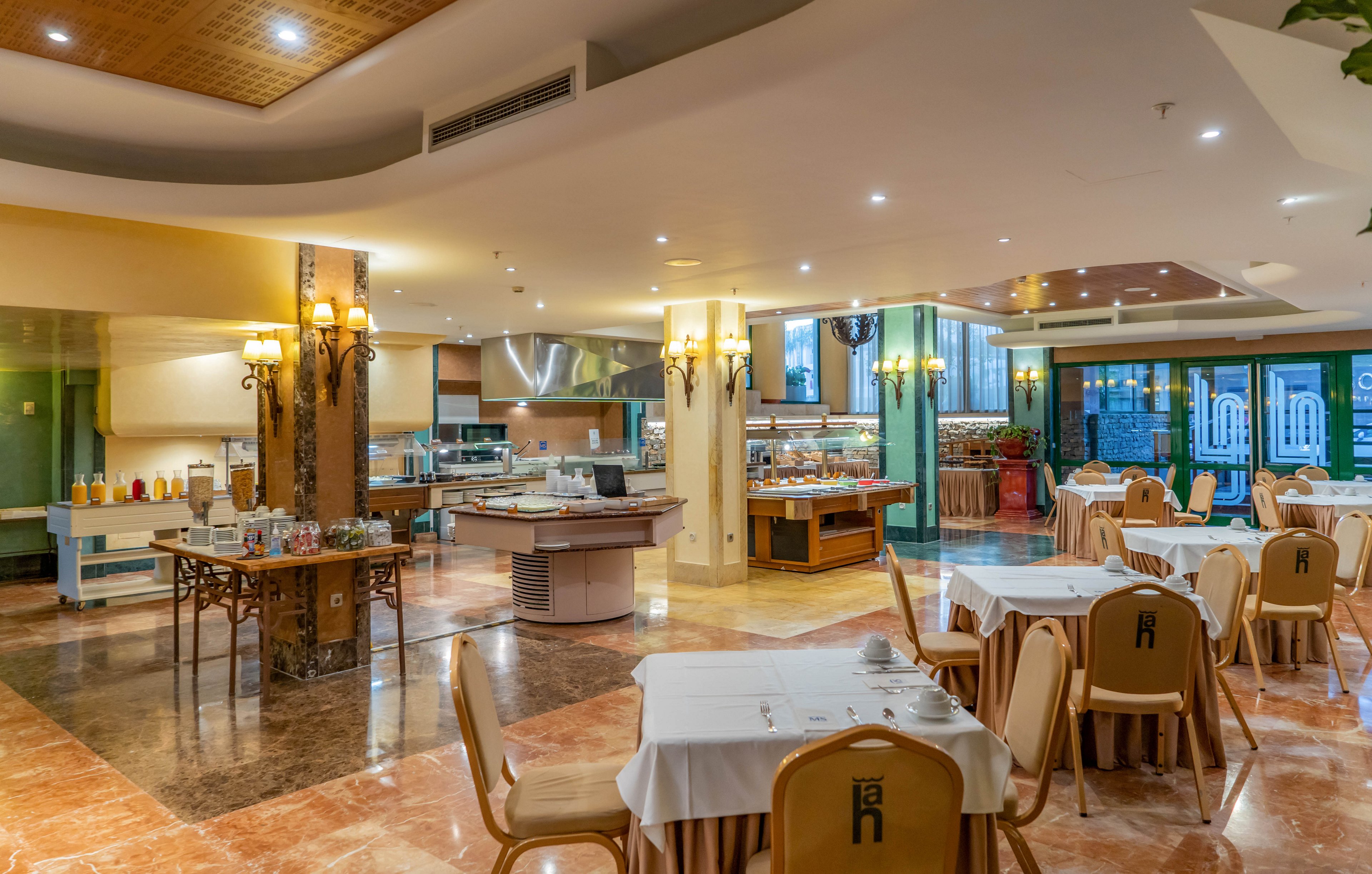 Bright, spacious restaurant interior featuring neatly arranged dining tables and chairs. A central buffet island and multiple food stations are visible in the background. Warm lighting, marble-style flooring, and decorative columns create an inviting atmosphere. Large windows on the right side suggest an evening or indoor resort setting.