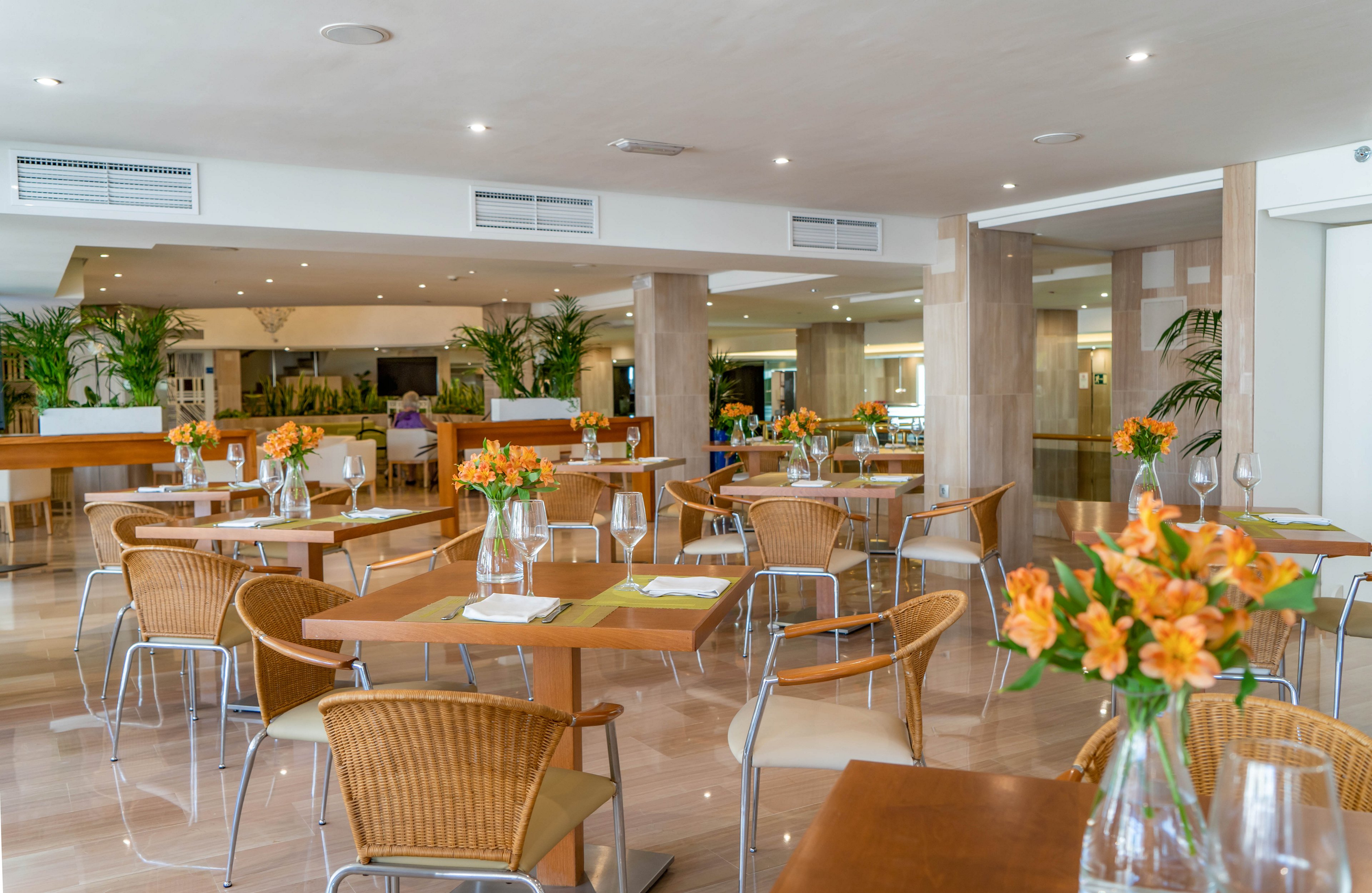 Spacious modern restaurant interior with neatly arranged wooden tables and wicker chairs. The room is brightly lit with recessed ceiling lights and large windows out of frame. Orange flowers in glass vases decorate the tables alongside empty wine glasses and folded napkins. Neutral tones, polished floors, and green plants create a clean, contemporary dining atmosphere.