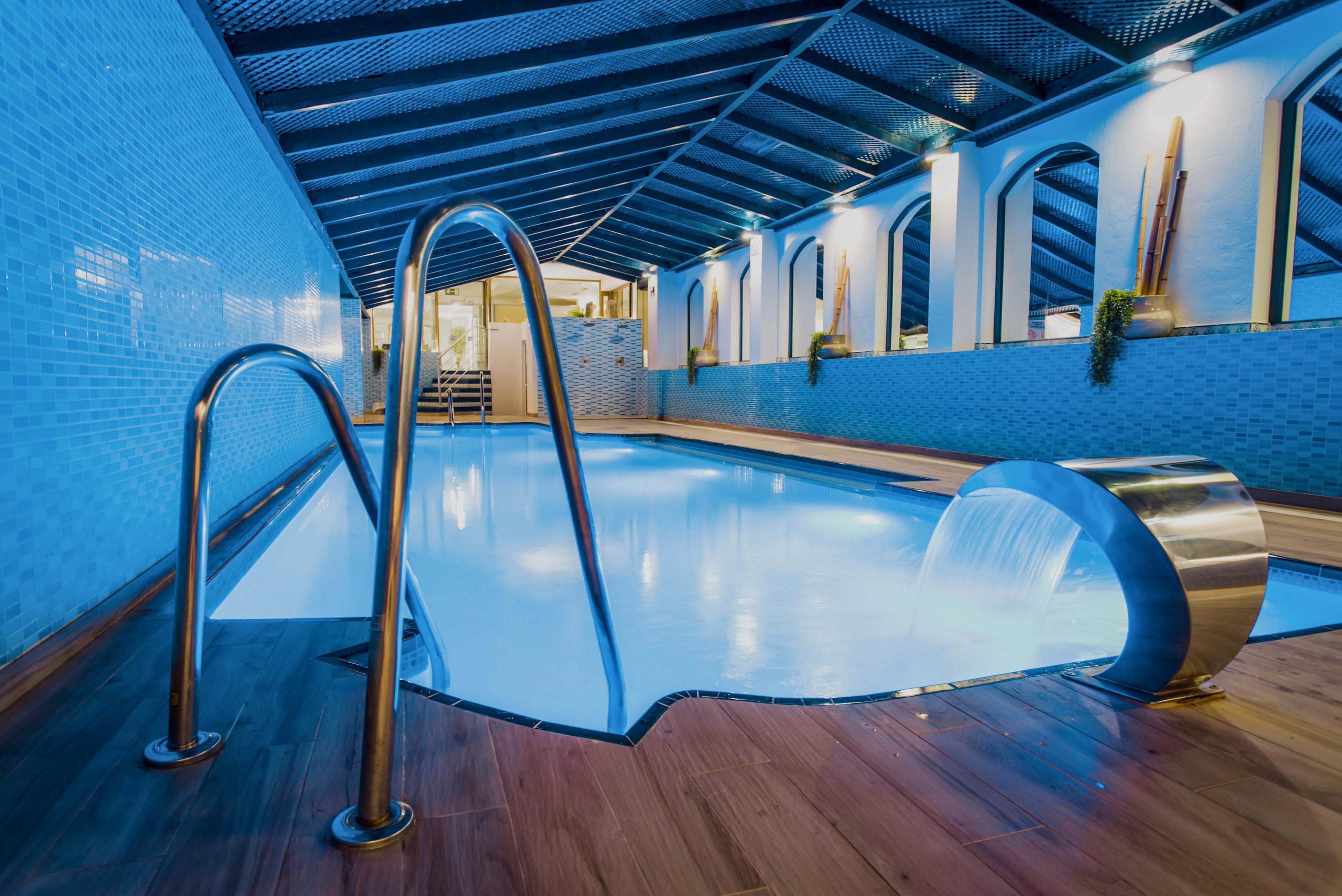An indoor swimming pool is shown under a modern, sloped roof with blue tiled walls. Stainless steel handrails lead into the clear water, which glows with cool blue lighting. A curved metal waterfall feature pours water into the pool near a wooden deck. The space appears clean, contemporary, and designed for relaxation or spa use.