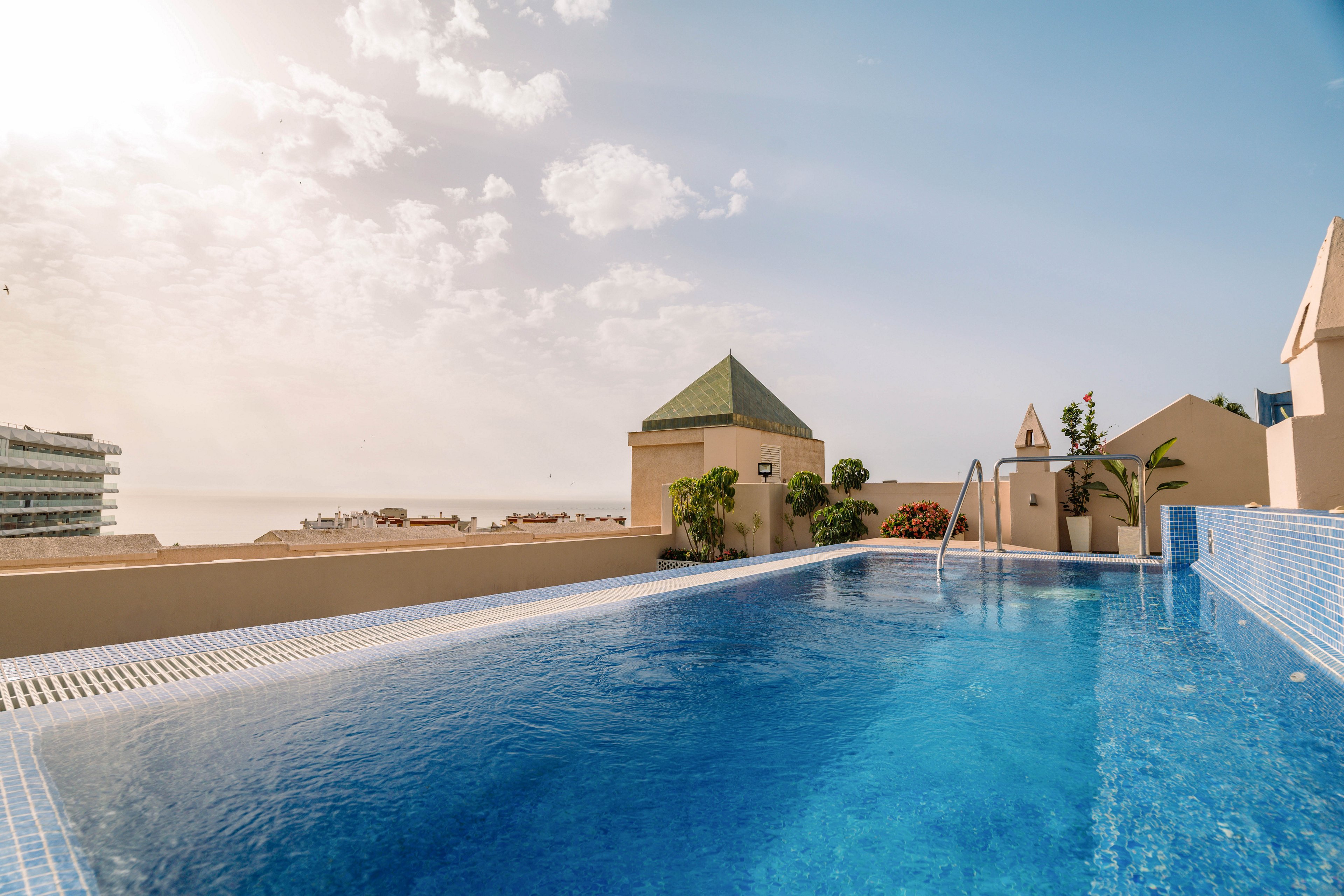 A serene rooftop infinity pool stretches toward the horizon under a hazy, sunlit sky. The pool is surrounded by low beige walls, potted greenery, and simple architectural structures. Metal handrails lead into the clear blue water, reflecting the warm afternoon light. In the distance, coastal buildings and the sea are softly visible, creating a relaxed vacation atmosphere.
