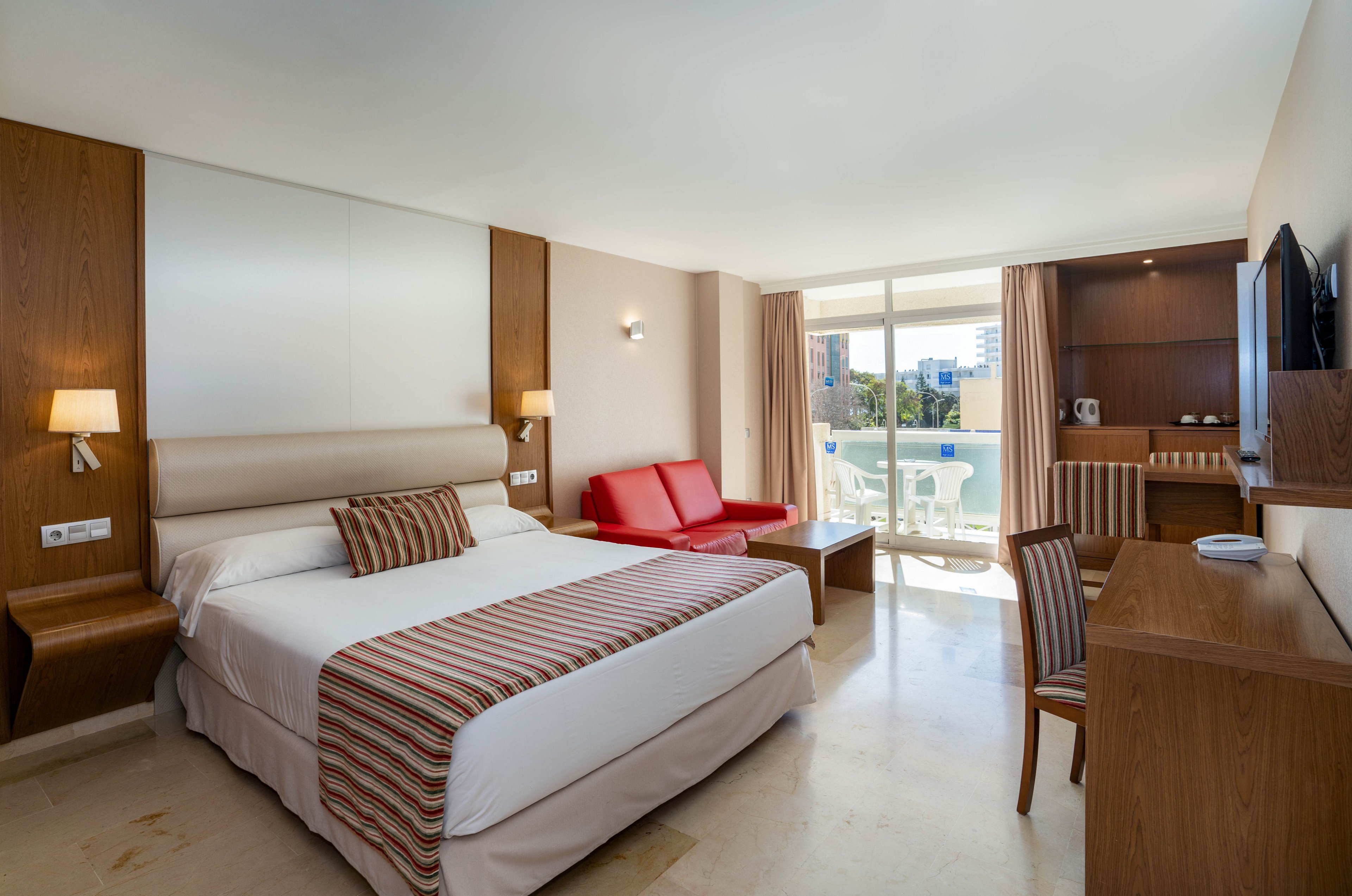 Spacious modern hotel room featuring a large double bed with striped bedding and coordinated pillows. The room includes wooden furniture, a desk with chair, wall-mounted TV, and bedside lamps. A red sofa seating area faces sliding glass doors that open to a bright balcony with outdoor chairs and a view of nearby buildings and greenery. Neutral walls and floors with warm wood accents create a clean, contemporary atmosphere.