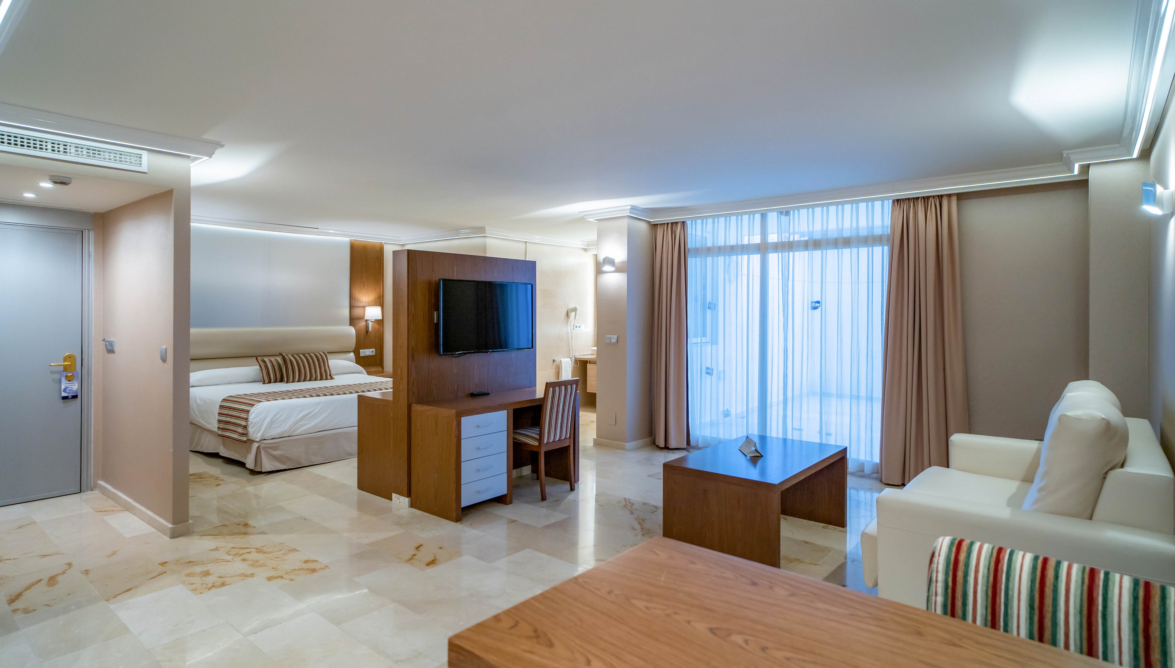 Bright, open-plan hotel suite featuring a large bed, sitting area, and work desk. A flat-screen TV is mounted on a central wooden partition facing both the bed and a sofa. Floor-to-ceiling windows with sheer curtains lead to an outdoor balcony, allowing natural light to fill the room. Neutral tones, tiled flooring, and simple contemporary furniture create a clean, relaxing atmosphere.
