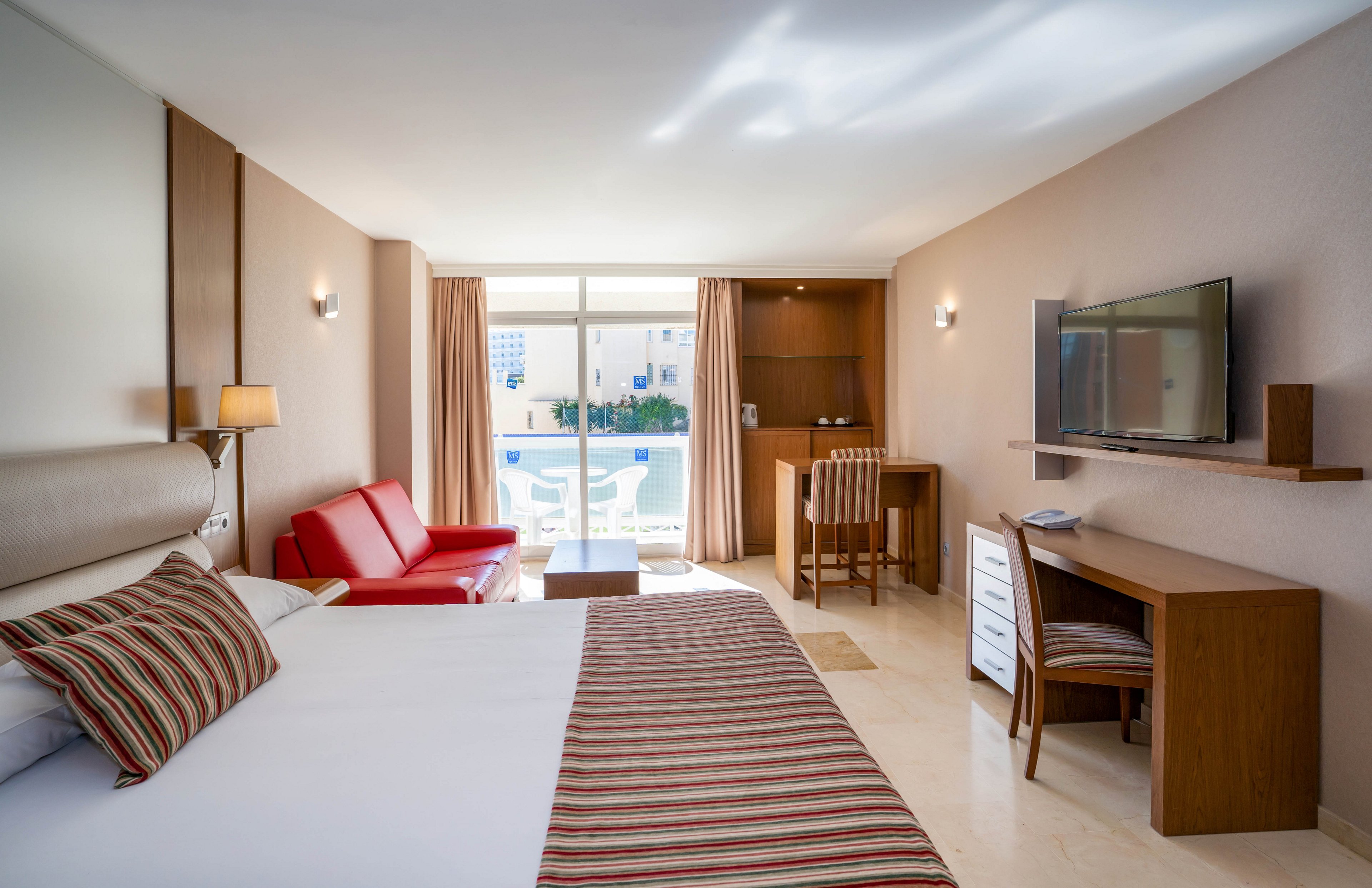 Bright contemporary hotel room featuring a large neatly made bed with striped pillows. The space includes a red sofa, wall-mounted flat-screen TV, and a wooden desk with chair. A small dining or work area stands near the sliding glass doors that open to a balcony overlooking a pool and other buildings. Neutral walls and light flooring create an airy, minimalist atmosphere with warm lighting.