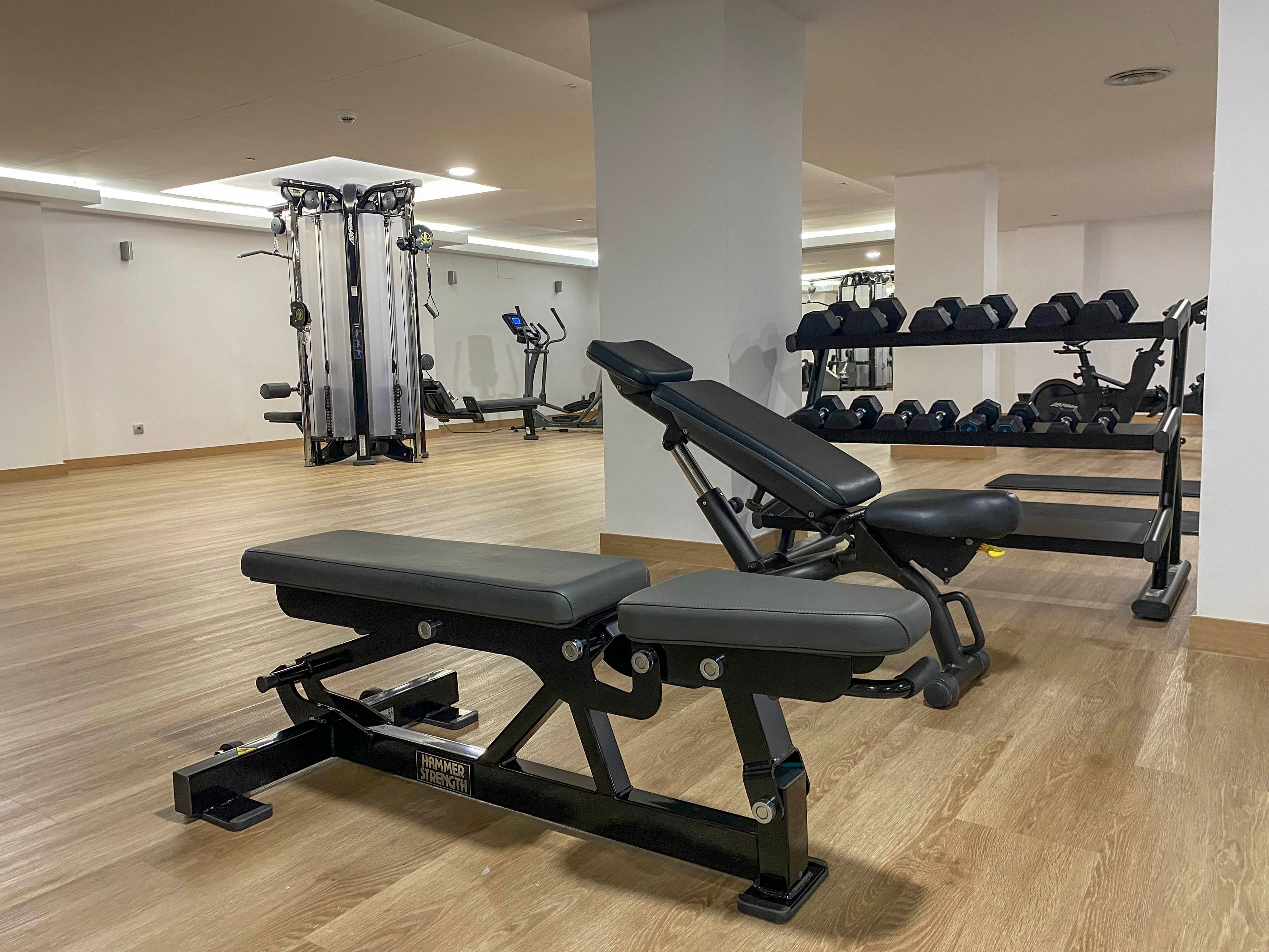 A clean, modern gym interior features adjustable weight benches in the foreground. In the background, a rack of dumbbells and a multi-station cable machine are visible. The room has light wood flooring, white walls, and bright overhead lighting, creating an open and minimalistic atmosphere. No people are present, emphasizing the equipment and spacious layout.