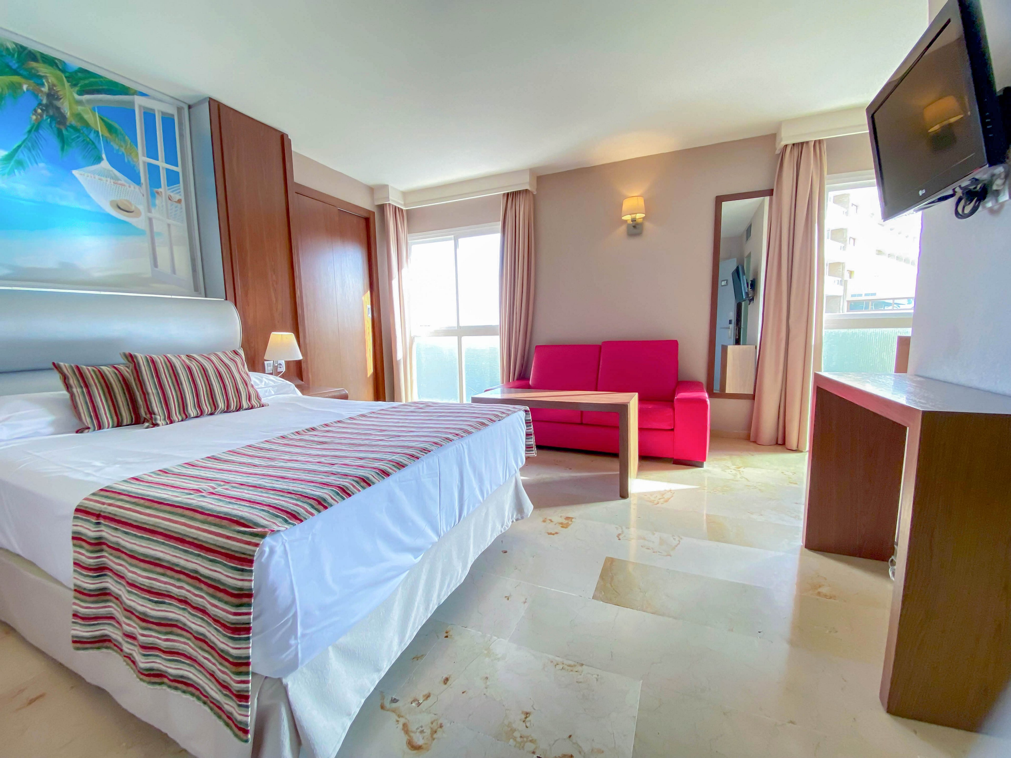 A spacious hotel room features a neatly made double bed with striped pillows and runner. Large windows with light curtains allow natural daylight to fill the space. A vivid pink sofa and wooden table sit near a full-length mirror, adding color contrast. A wall-mounted television and beach-themed artwork complete the modern, inviting interior.