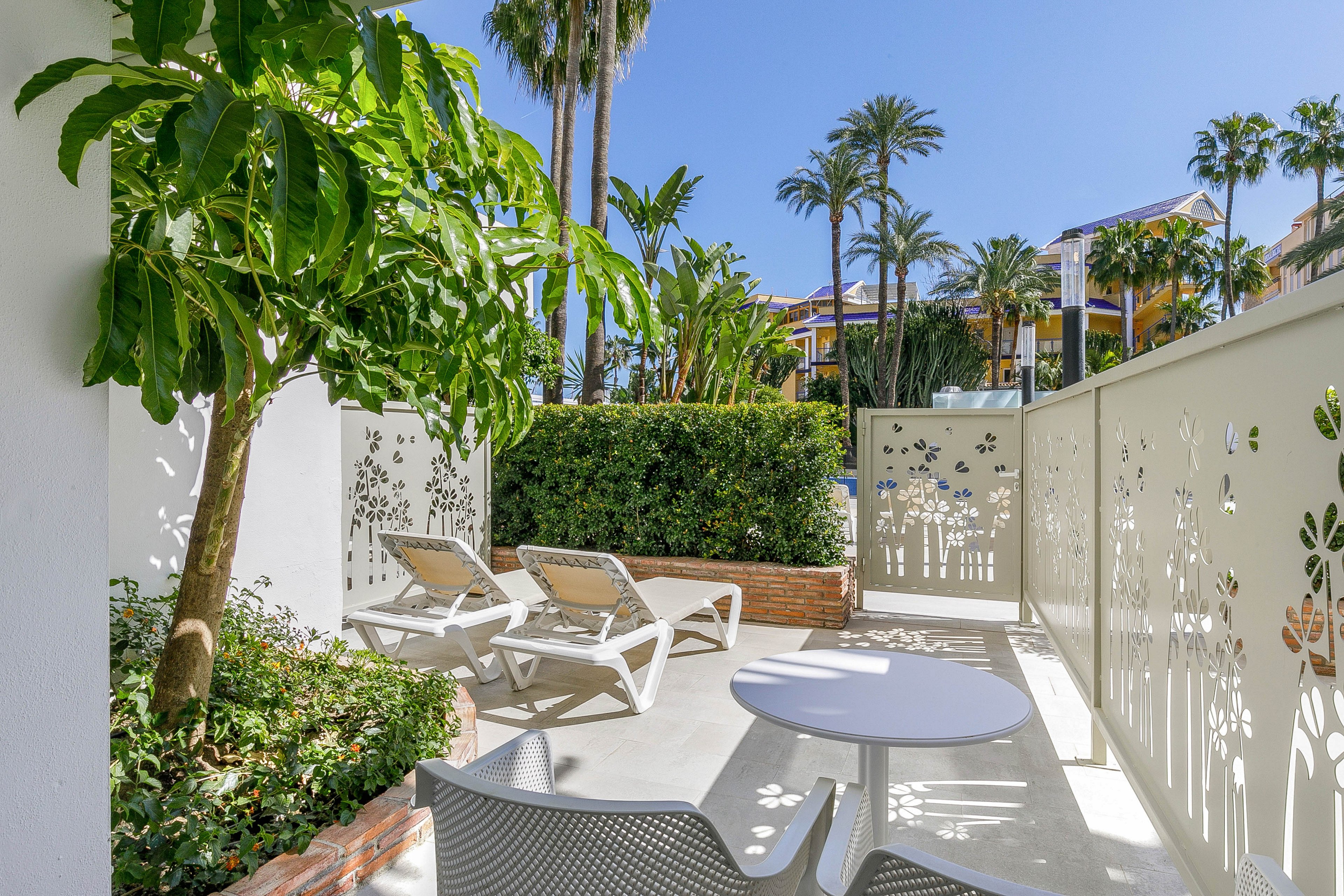 A bright outdoor patio features two white lounge chairs and a small round table under clear blue skies. Decorative perforated panels with floral patterns create a sense of privacy while still letting sunlight through. Lush green plants and palm trees surround the space, suggesting a resort or vacation setting. The overall atmosphere is relaxed and inviting, ideal for sunbathing or quiet outdoor leisure.