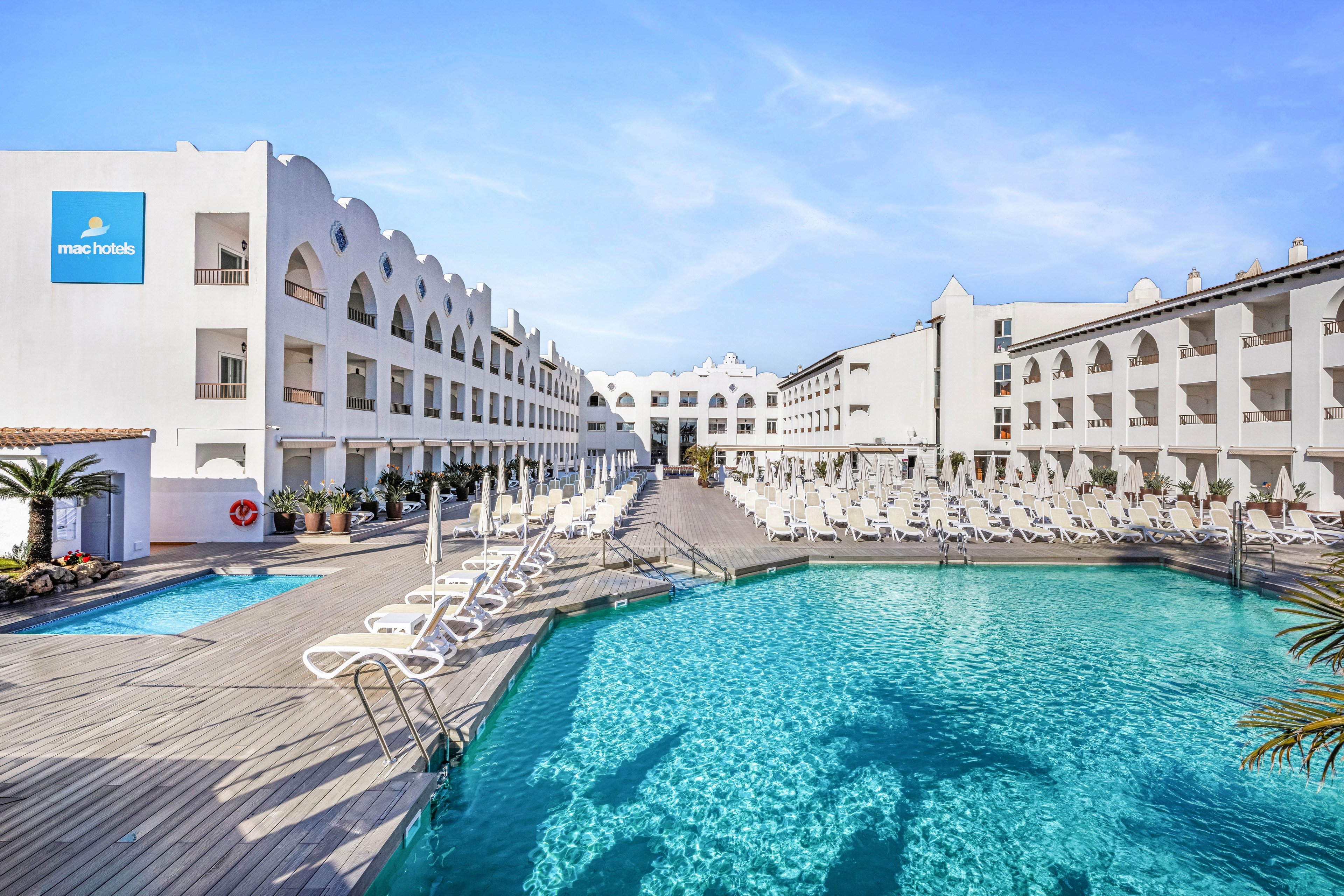 A large outdoor swimming pool is surrounded by white hotel buildings with arched balconies. Numerous sun loungers and umbrellas line the wooden deck, suggesting a relaxing vacation setting. The clear blue water of the pool reflects the bright sky, creating a vibrant and inviting atmosphere. Palm trees and clean architectural lines add to the modern Mediterranean resort style.