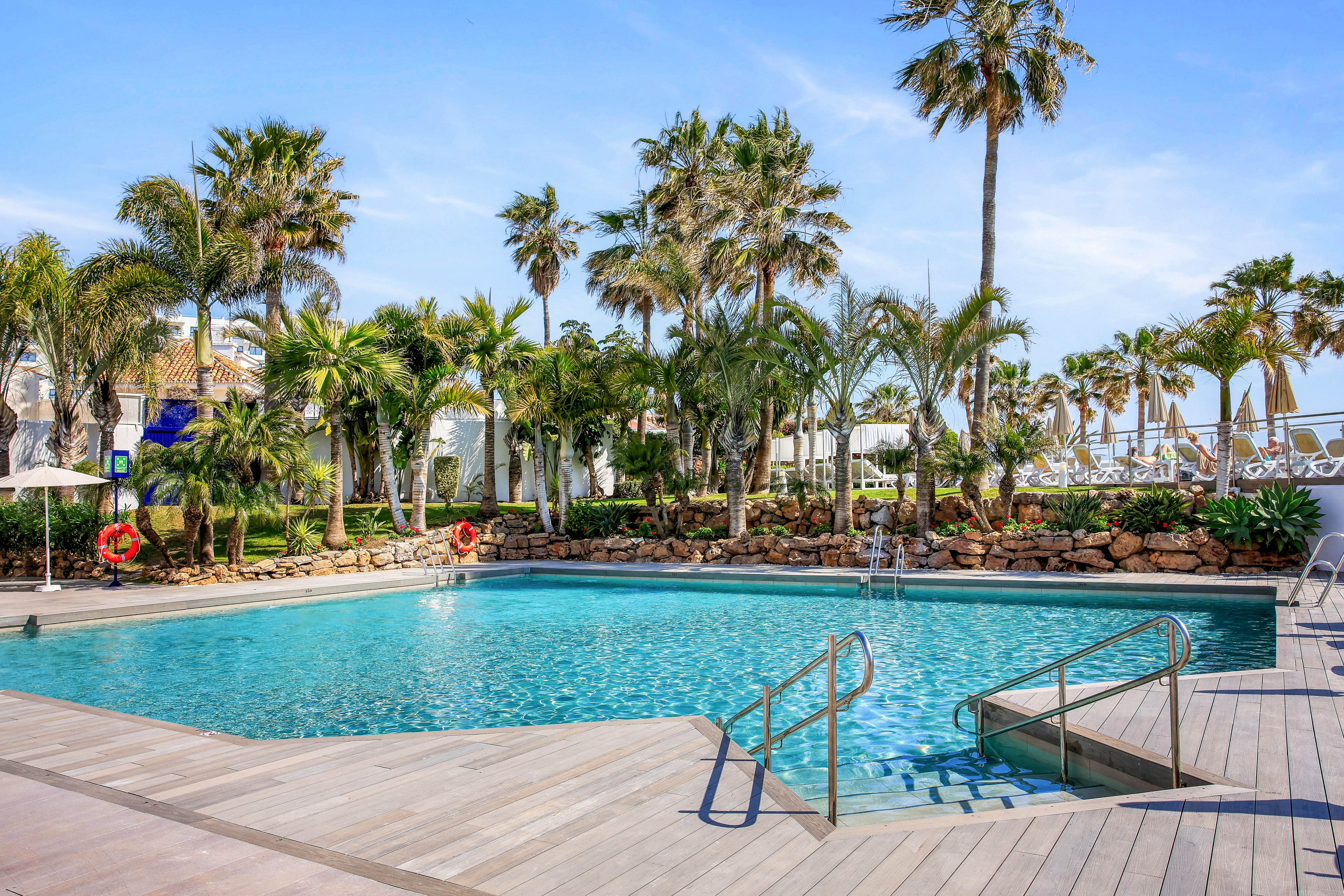 An outdoor swimming pool is surrounded by tall palm trees and lush greenery at a coastal resort. The clear blue water reflects the bright sky, while metal handrails lead into the pool from a wooden deck. Life rings hang along the stone wall, and white buildings and structures are visible in the background. The scene suggests a relaxing vacation atmosphere in warm, sunny weather.