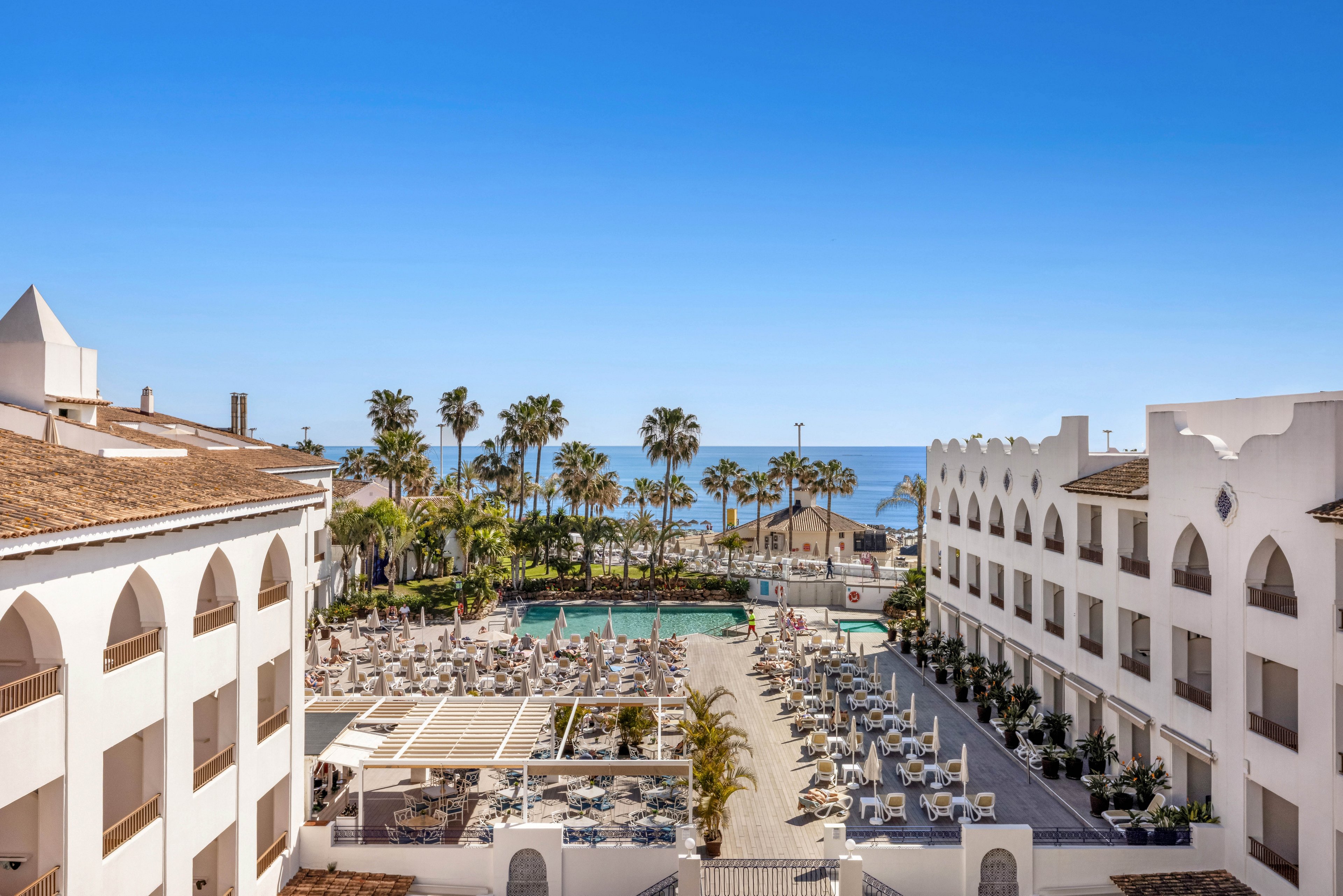 A coastal resort complex features white buildings framing a central outdoor pool area. Numerous sun loungers and umbrellas are arranged around the pool, with palm trees adding a tropical touch. The scene opens toward a calm blue sea under a clear sky, suggesting a warm, sunny vacation destination. Architectural arches and balconies line the buildings, emphasizing a clean, Mediterranean-inspired style.
