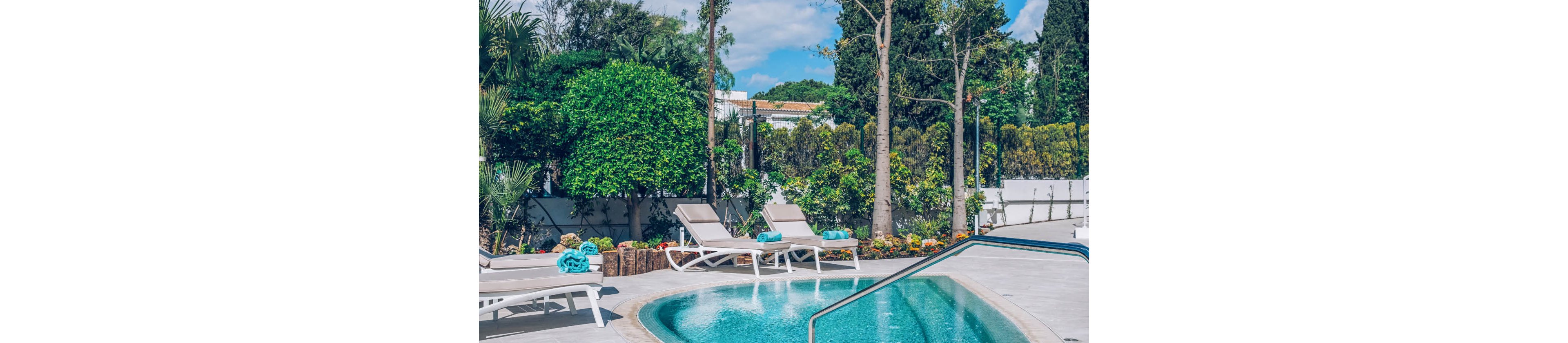 An inviting outdoor swimming pool is surrounded by several white lounge chairs and bright turquoise towels. The setting appears to be a landscaped garden with lush green trees, shrubs, and colorful flowers along the poolside. A metal handrail leads into the clear blue water, and white buildings with tiled roofs are visible in the background. The scene suggests a relaxing, sunny day in a private villa or resort.