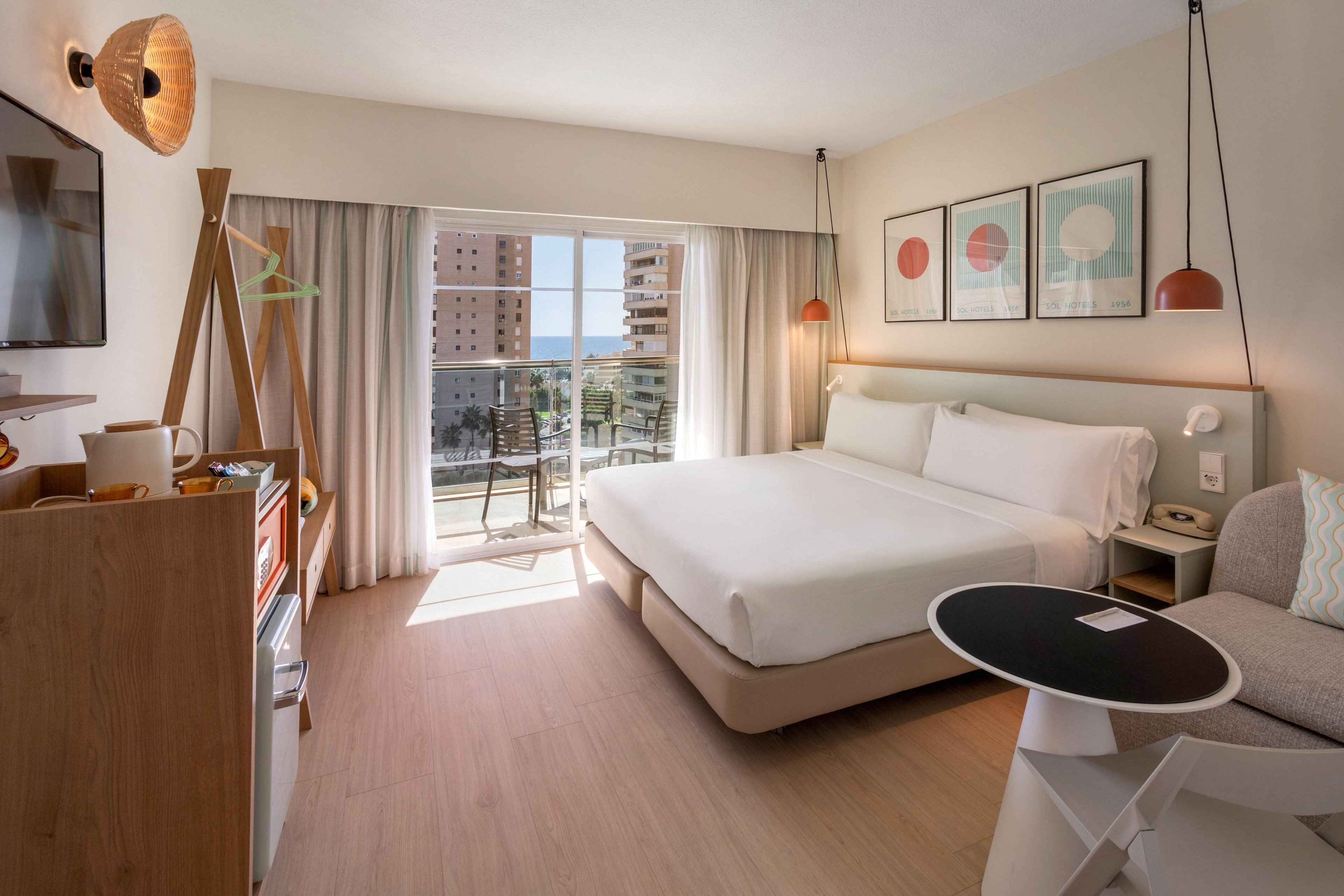 Bright contemporary hotel room featuring a large neatly made bed and minimalist furnishings. A sliding glass door opens to a balcony with chairs, overlooking tall buildings and a distant ocean view. Soft neutral tones, wooden flooring, and simple wall art create a calm, inviting atmosphere. Natural daylight fills the space, highlighting the compact workspace, seating area, and small kitchenette.