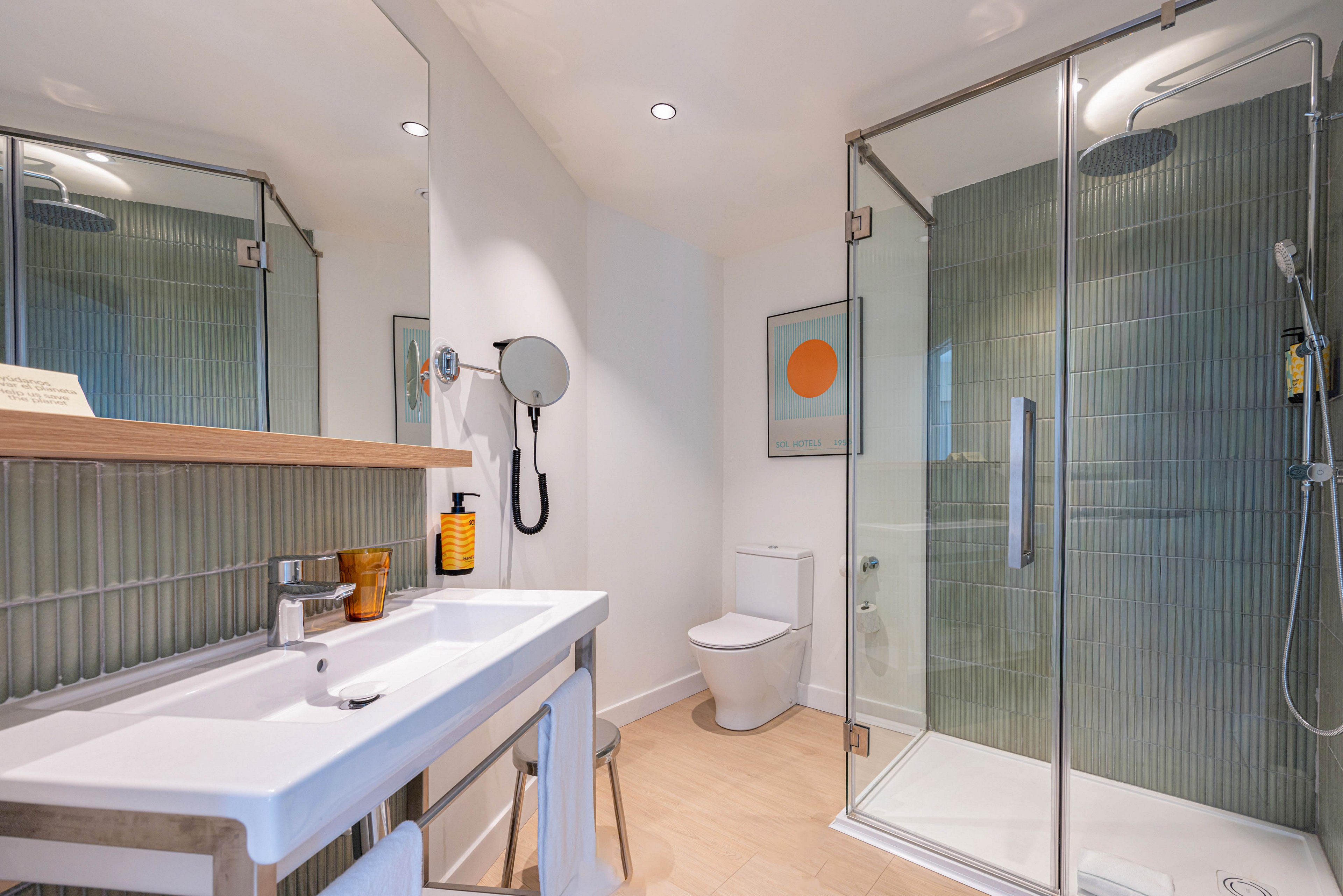 Contemporary bathroom interior featuring a large glass-enclosed shower and a white ceramic toilet. A wide rectangular sink with a sleek countertop holds soap dispensers and small accessories. Vertical green wall tiles and light wood flooring create a clean, modern look. A round wall mirror, recessed ceiling lights, and a colorful abstract artwork add stylish finishing touches.