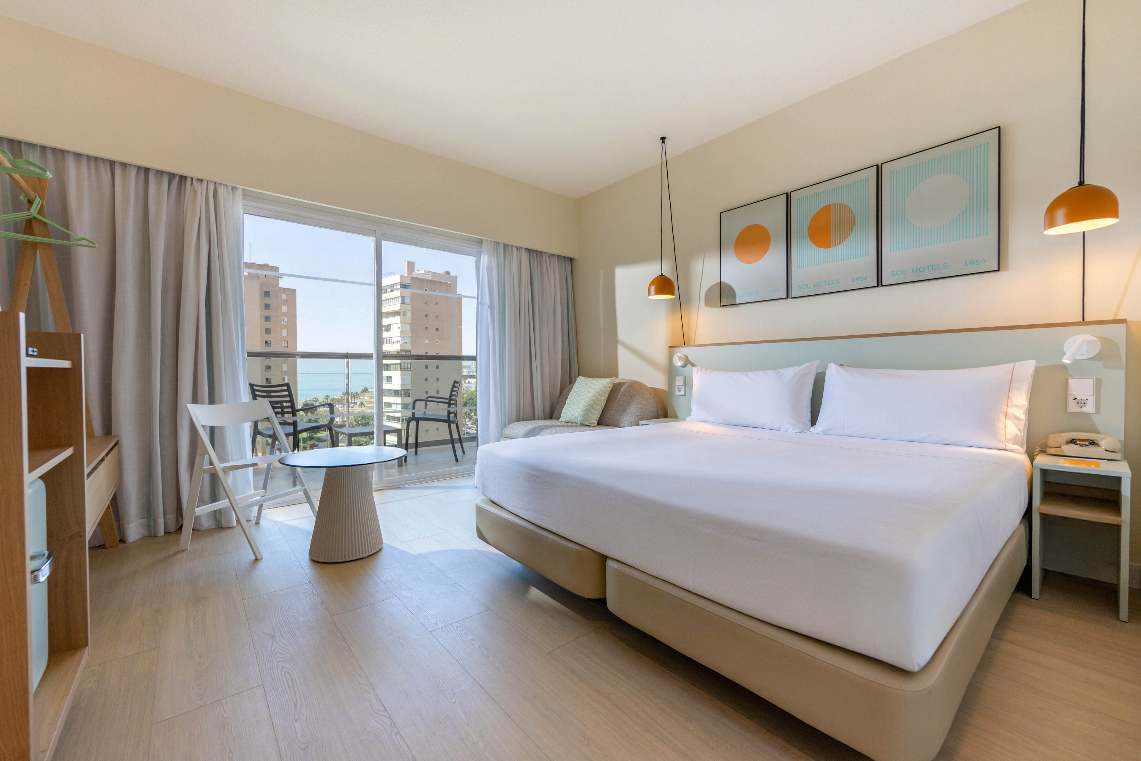 Bright contemporary hotel room featuring a large neatly made bed and minimalist furniture. Floor-to-ceiling windows open onto a balcony with chairs and a table, overlooking city buildings and a distant water view. Soft neutral tones are accented by pastel artwork and warm pendant lighting. The space appears clean, inviting, and designed for comfortable urban accommodation.